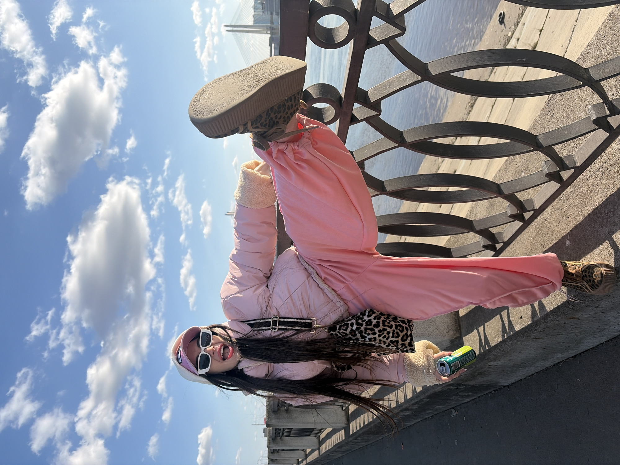 A female individual is depicted in a dynamic pose on a concrete embankment alongside a wide body of water in Saint Petersburg, Russia, with a prominent cable-stayed bridge, identifiable as part of the Western High-Speed Diameter, visible in the distant background. The subject, a young adult with long dark hair, wears white rectangular sunglasses, a white beanie over a pink baseball cap, a light pink quilted puffer jacket with light-colored fleece-lined cuffs, and matching light pink wide-leg pants. She is wearing leopard-print platform shoes/boots and carries a leopard-print shoulder bag with a black and white striped strap. Her right leg is elevated, with her leopard-print shoe resting on the upper rail of the dark metal embankment railing, which features repeated oval and X-shaped decorative elements. Her left foot remains on the paved surface of the embankment. She holds a green and white beverage can in her right hand. Her mouth is open wide, revealing teeth and tongue, suggesting an expression of excitement or laughter. The scene is illuminated by bright sunlight under a clear blue sky with scattered white cumulus clouds. Below the railing, a concrete slope descends to the water's edge.