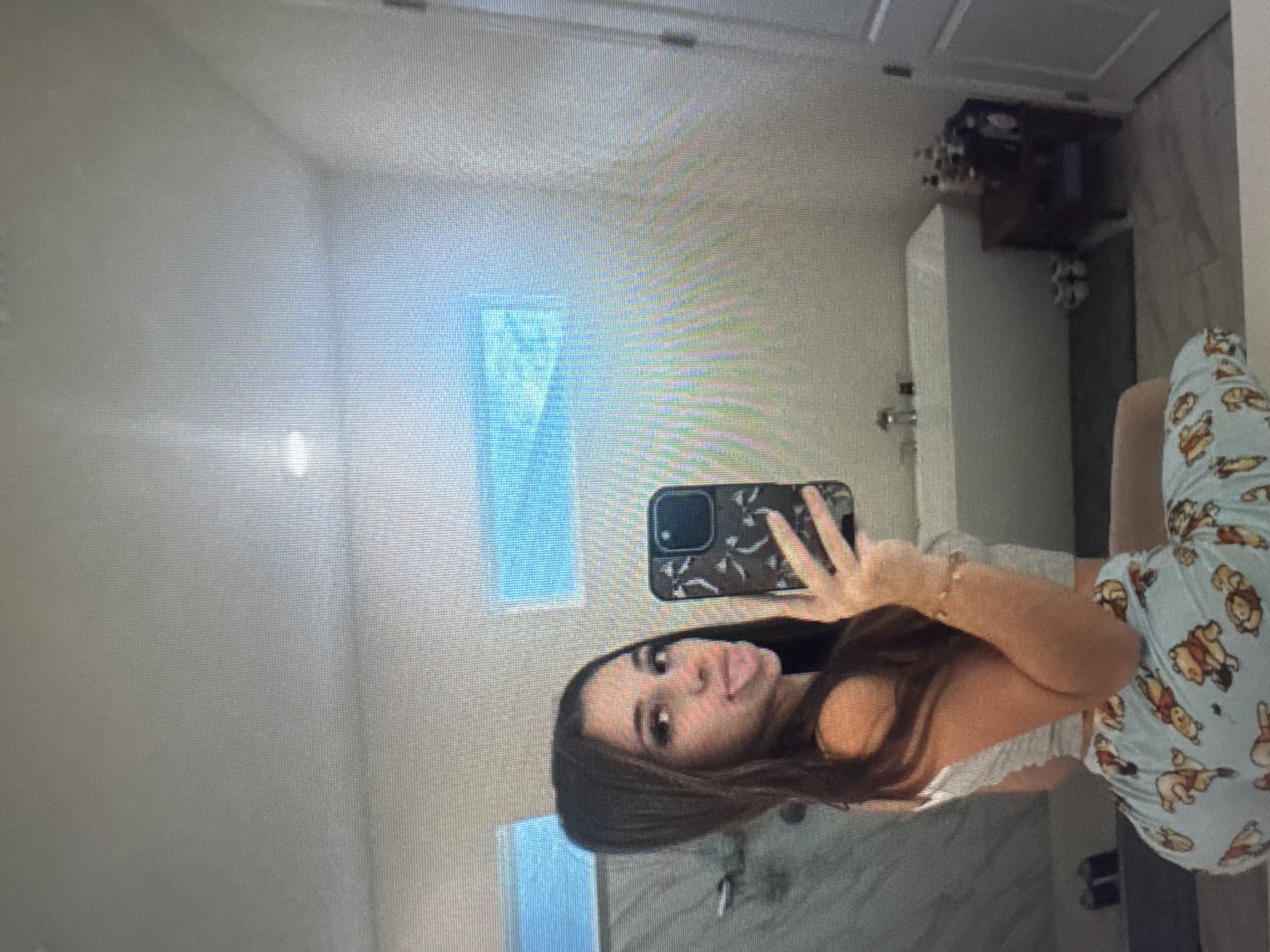 An image, rotated 90 degrees counter-clockwise, depicts a young woman taking a mirror selfie in what appears to be a bathroom. The woman, with long dark brown hair, is positioned centrally, looking directly at her smartphone, which she holds up with her right hand, making a peace sign gesture. Her tongue is slightly sticking out, conveying a playful or casual expression. She is wearing a light-colored crop top and light blue pajama pants adorned with cartoon bear characters, reminiscent of Winnie the Pooh. A subtle bracelet is visible on her right wrist.
The background of the selfie, reflected in the mirror, shows a clean, light-colored bathroom with what appears to be white or light grey tiled walls. A sink and faucet are visible beneath the mirror, and a dark wooden cabinet or vanity is partially visible to the right. Bright reflections or actual windows in the background show a light blue tint, indicating daylight. No specific cues within the image directly suggest the location is Varna, Bulgaria. No legible text is visible in the image.