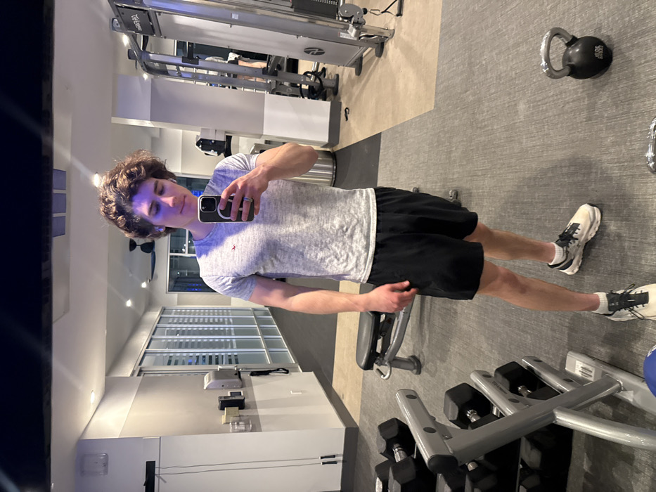 A young adult male, exhibiting curly brown hair and wearing white wireless earbuds, stands in a fitness center located in Los Angeles, United States of America. He is wearing a light gray t-shirt with a small red moose logo on the left chest, black athletic shorts, white socks, and white athletic shoes. The male is holding a black smartphone with a clear magnetic case in his right hand, reflecting his image in a mirror.
The environment is an indoor gym with a gray rubber mat floor, transitioning to a lighter tan section in the background. Walls are light gray and beige. Overhead recessed lighting illuminates the space, casting a slight bluish tint. Exercise equipment visible includes a two-tier metal dumbbell rack holding multiple black hex dumbbells (e.g., 20 lbs, 25 lbs), a black adjustable weight bench behind the subject, and a black kettlebell (approximately 30 lbs) on the floor to the subject's right. Further in the background, various weight machines, cable apparatuses, and a metal trash receptacle are present. A blue medicine ball is partially visible on the floor in the lower left. A row of large windows with horizontal opaque coverings line a wall in the background, behind which a dark exterior is visible. A wall-mounted hand sanitizer dispenser and a black jump rope are visible on the wall to the left. Another individual is partially visible operating a piece of equipment in the far right background.