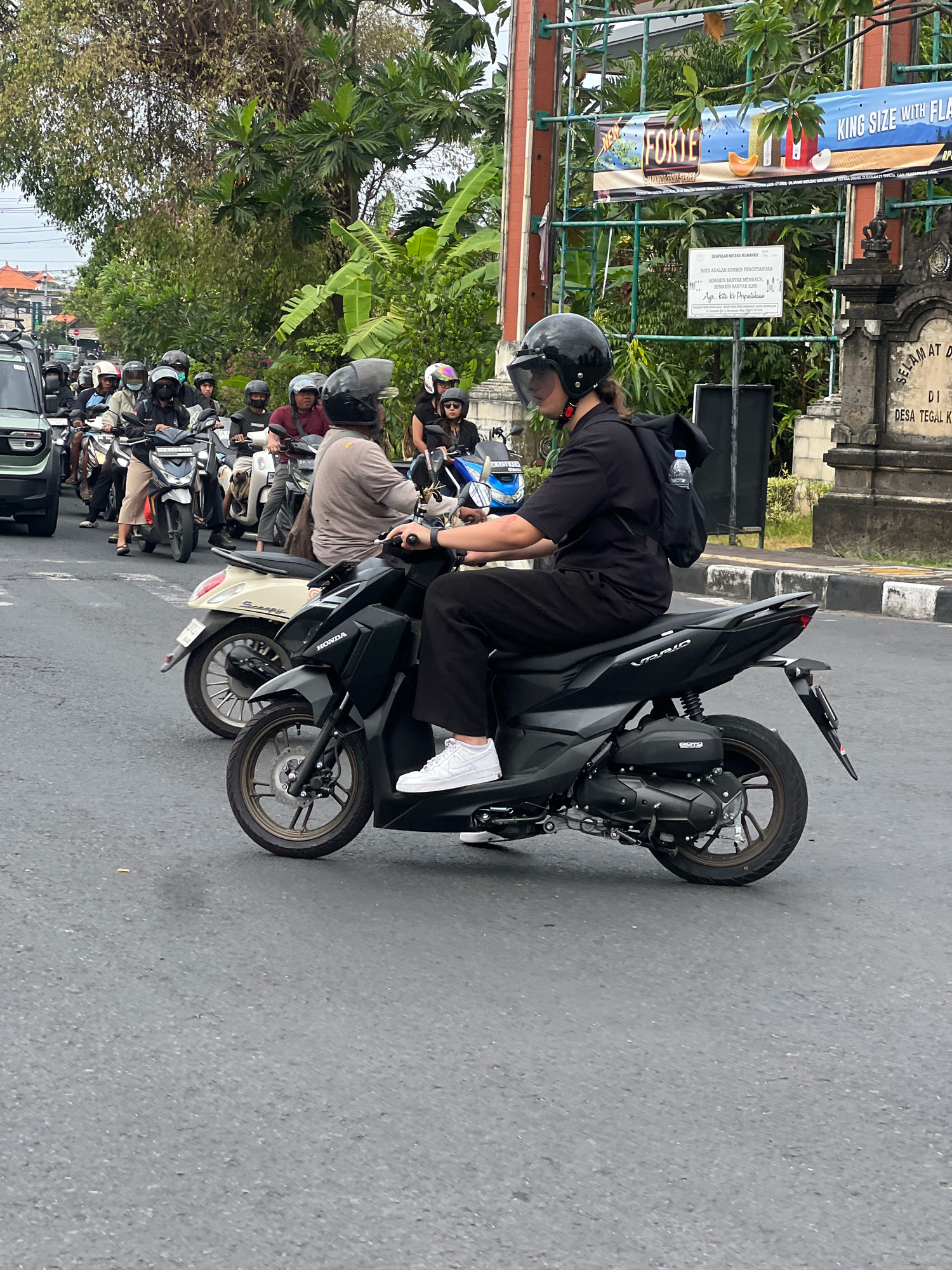 This is a street-level, eye-level, medium shot of a traffic jam on a paved road in Denpasar, Indonesia. The foreground is dominated by a black Honda Vario scooter, ridden by a person wearing a black helmet, black short-sleeved shirt, black pants, and white sneakers. A bottle of water is clipped to a black backpack worn by the rider. Behind this scooter, other scooters and motorcycles are visible, with riders and passengers also wearing helmets. In the background, there are trees, a fence, a gate with inscriptions, and a sign with text and images. The lighting suggests an overcast day. The overall scene depicts typical urban traffic in Denpasar.