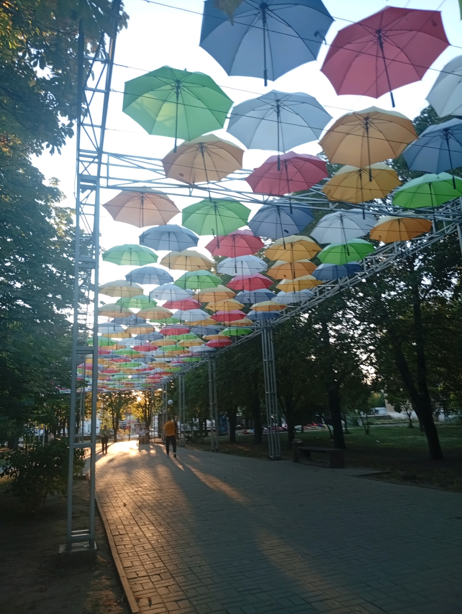 A wide-angle, low-angle shot captures an outdoor scene in Pokrov, Ukraine, featuring a pathway lined with trees and a canopy of colorful umbrellas suspended overhead. The umbrellas, in various shades of blue, green, red, yellow, and white, are strung across a metal frame structure, creating a vibrant, whimsical atmosphere. Sunlight streams through the umbrellas, casting dappled shadows on the brick-paved pathway. Two figures, silhouetted against the bright light, are walking along the path. The figure in the foreground is wearing dark pants and a yellow shirt, while the figure behind them is wearing dark clothing. Trees with lush green foliage flank the pathway, and in the background, more trees and buildings are visible. The overall impression is one of a lively public space decorated for an event or festival.