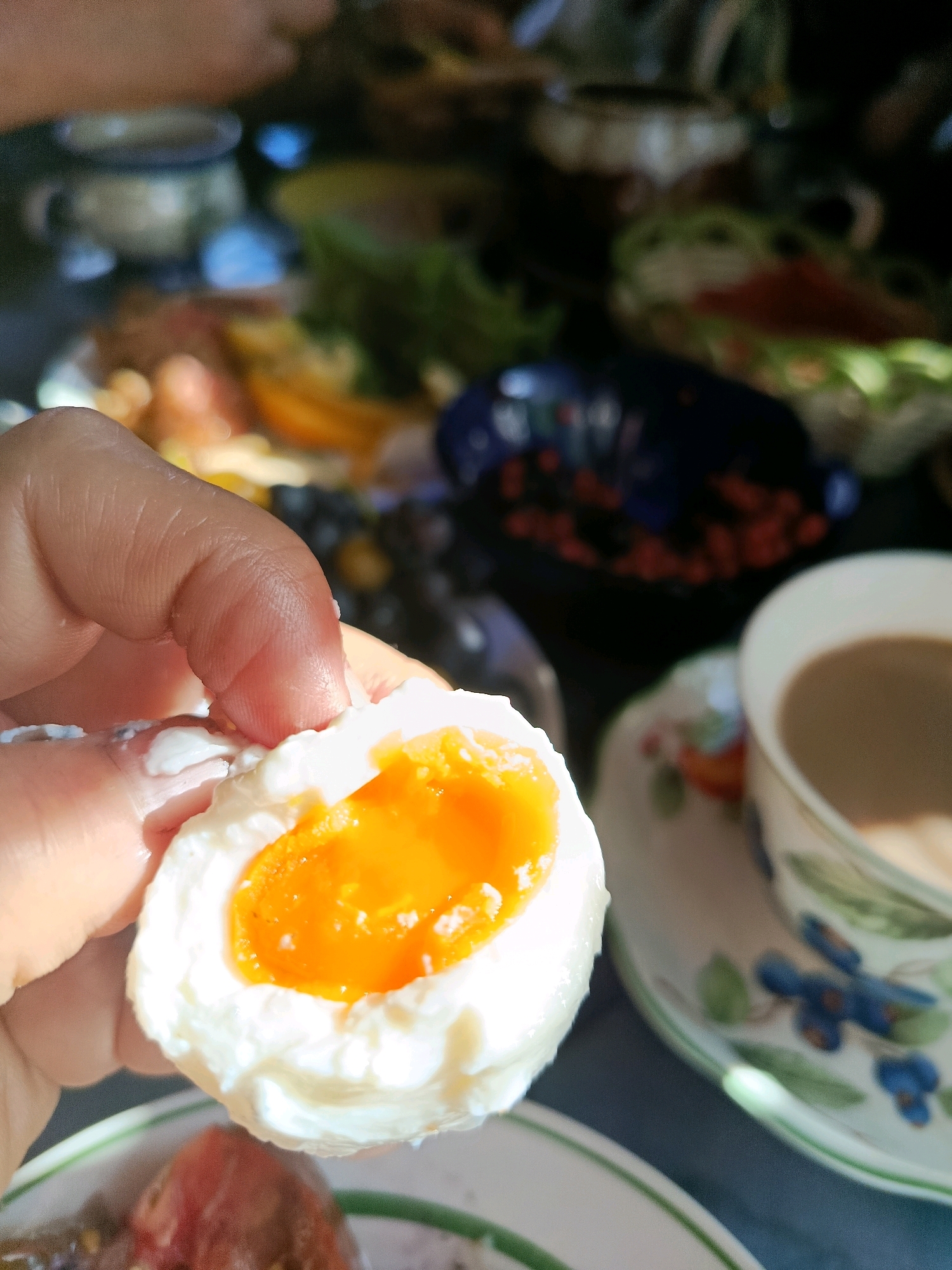 In an outdoor setting, a hand holds a half-eaten soft-boiled egg. The egg white is firm and white, while the yolk is a bright, runny orange, dripping slightly onto the fingers holding it. In the background, a cup of coffee sits on a saucer with a floral design. Further back, other food items and dishes are visible, suggesting a meal setting. The lighting indicates it is daytime. The scene is foreshortened from a high-angle perspective.