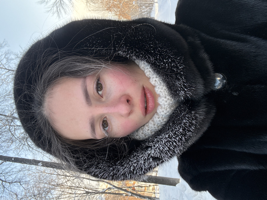 A female subject, appearing to be approximately 20-30 years old, is depicted in a close-up, head-and-shoulders portrait against a winter outdoor backdrop in Yekaterinburg, Russia. She looks directly at the viewer with a neutral expression. Her skin exhibits erythema on the malar regions and nasal tip, consistent with cold exposure. Frost accretion is visible on her eyelashes, particularly the left, and a minor deposit on the nasal tip. Her lips are slightly parted. Dark brown hair is partially visible around her face, some strands showing frost or grey coloration. She wears a dark, likely black, fur coat with a prominent hood. The hood is lined with contrasting light-tipped fur, which also shows evidence of frost. Around her neck is a light-colored, knitted neck warmer with a visible textured pattern. A large, dark, domed button is present on the coat's front closure. The background consists of a snowy landscape with bare, snow-dusted deciduous trees under a clear, light blue sky. A multi-story building is partially visible in the distant left background, indicating an urban or park setting. The lighting suggests a bright, cold day.
