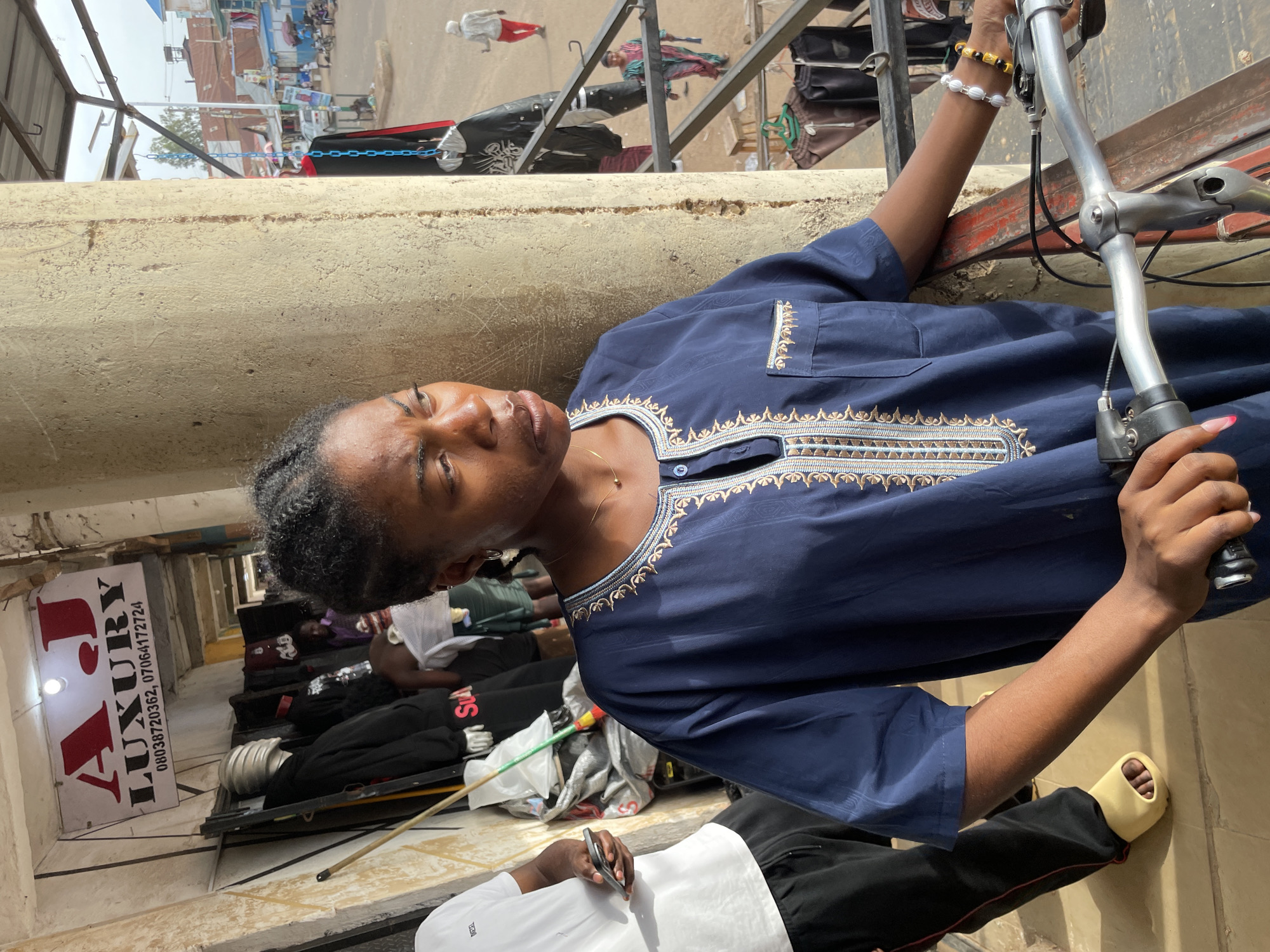 A young woman with braided hair stands in front of a store labeled "A.J. LUXURY" in a bustling market. She is wearing a dark blue garment with ornate gold embroidery around the neckline and a breast pocket. Her right arm rests on a bicycle handlebar, and her left hand is on the brake lever. To her left, a person in a white t-shirt and black pants holds a smartphone. Behind the woman, mannequins display clothing, and various items are hung for sale. The market appears to be outdoors, with a street visible in the background, featuring shops, people, and vehicles. The lighting suggests it is daytime.