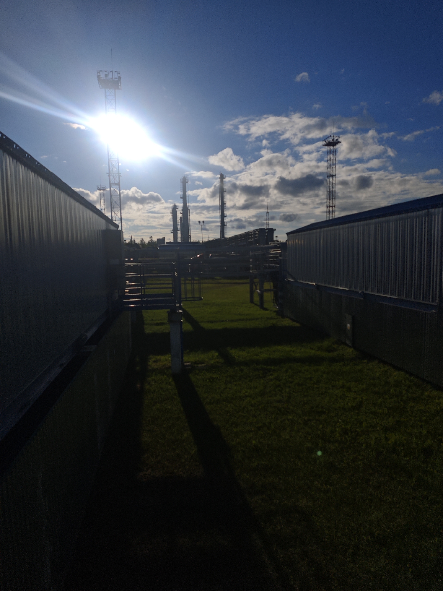 The image depicts an industrial site in what may be Unknown, Russia. The scene is set between two long, rectangular metal buildings with corrugated siding. An area of grass is visible between the buildings, with shadows cast from various structures. In the background, several industrial towers and pipes are visible against a partly cloudy sky. The sun is bright and slightly obscured by one of the towers, creating a lens flare effect. There are no people visible in the image. The overall environment suggests an outdoor industrial area.