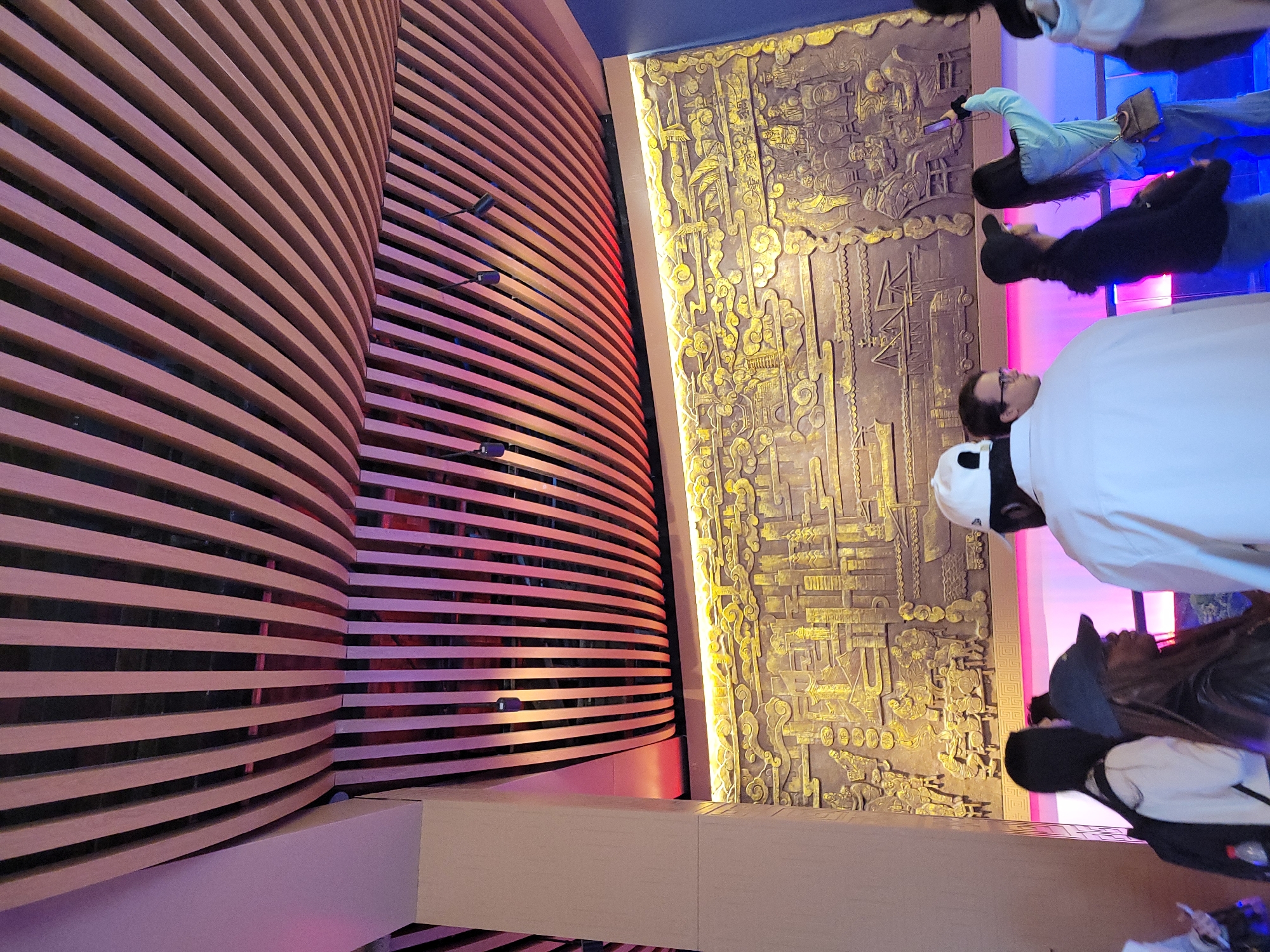 The image shows an interior view of a building in Hechuan, China. A curved wooden wall made of horizontal slats is prominently featured on the left. To the right is a decorated wall panel with gold and brown coloring, depicting a historical scene or figures. Several people are standing in front of the wall panel, appearing to be observing it. One person is wearing a white cap and a white shirt. The overall lighting in the area is provided by colored lights including blue and pink.