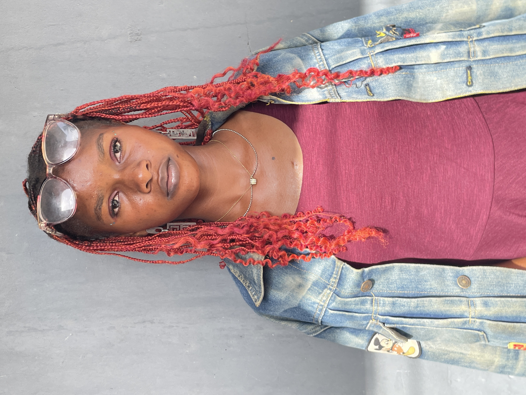 The image, rotated 90 degrees clockwise, presents a portrait of a young Black woman against a plain grey background. She has long, vibrant red braided hair, with some sections styled into distinct, tight curls. Her clear-framed glasses are pushed up onto her forehead, resting on her hair. She looks directly at the camera with a neutral expression. She is dressed in a fitted burgundy or maroon top, layered under a light blue denim jacket. The jacket features subtle distressing and patches, notably a Mickey Mouse patch visible on the lower front. A delicate gold-toned necklace with a small rectangular charm is worn around her neck, and she has geometric-designed earrings. The setting is Badagry, Nigeria.