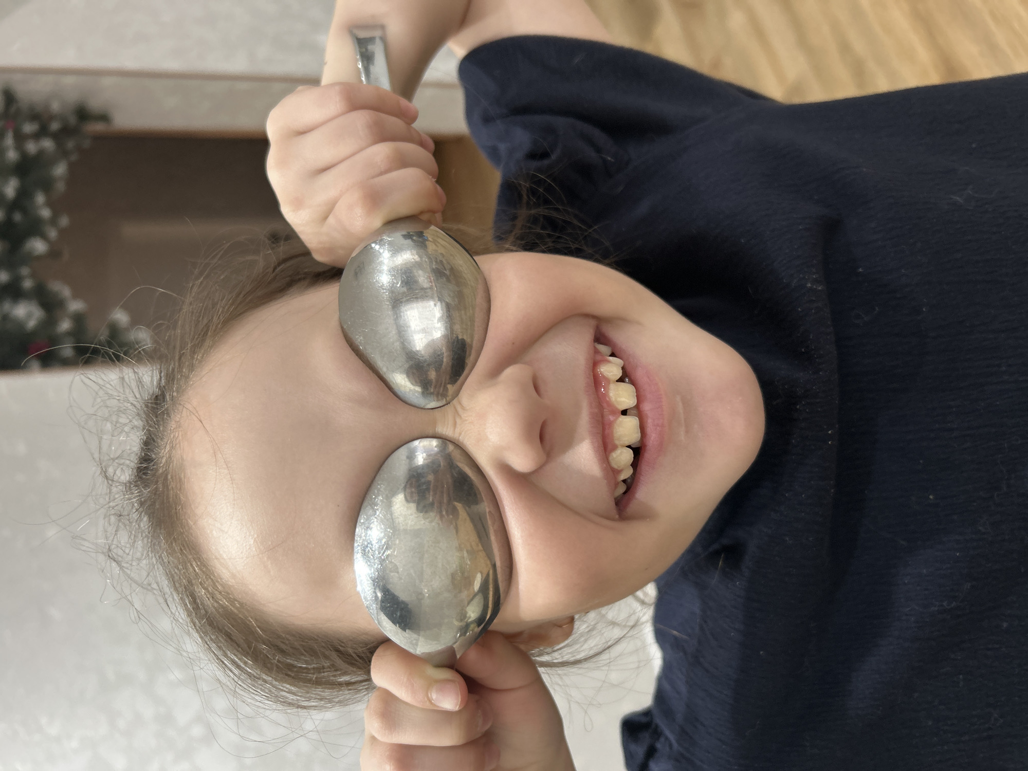A child with fair skin and light brown hair is holding two silver spoons to their eyes and smiling. They are wearing a dark blue shirt. The background includes a white wall, a wooden surface, and a blurry image of what appears to be a decorated Christmas tree. There is no clear indication of Chaikovsky, Russia.