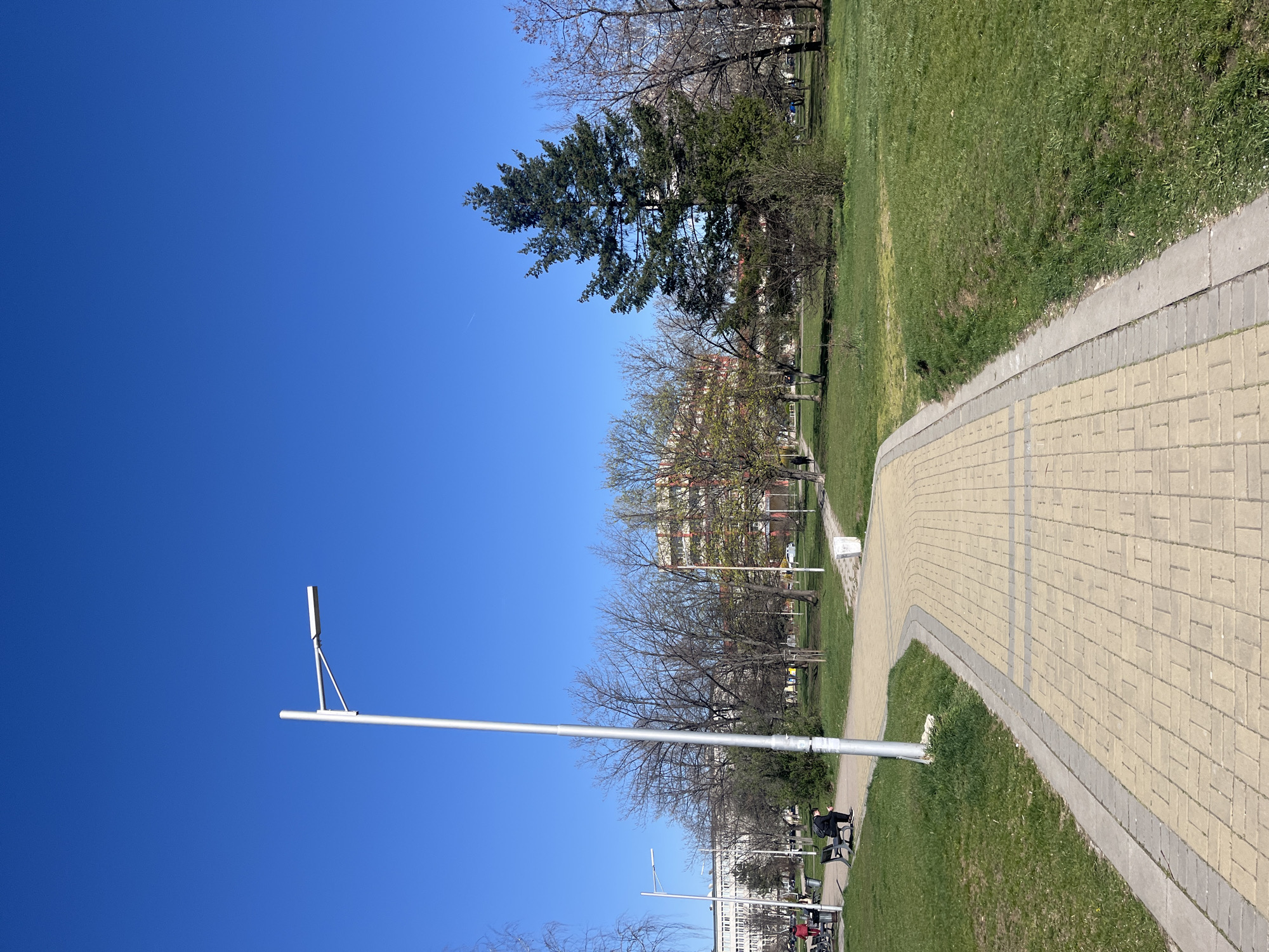 An outdoor scene shows a park on a clear day in Sofia, Bulgaria. A paved walkway curves through green grass and trees. A tall light pole stands near the path, extending into the blue sky. People are seated on benches near the walkway. Buildings are visible in the background.