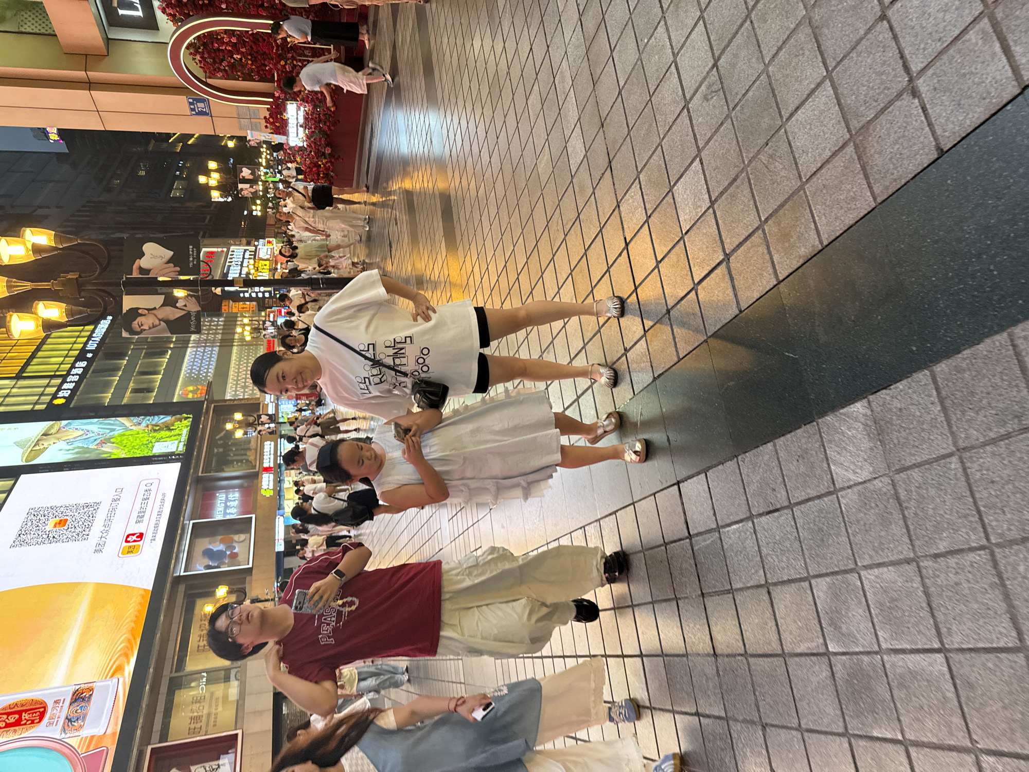 The image depicts a bustling street scene in the Yuzhong District of China. A group of people are walking on a tiled pedestrian walkway. In the foreground, a young girl in a light blue dress is looking at her phone. To her left, a woman in a white t-shirt and black shorts also appears to be looking at her phone. Further to the left, a young man wearing glasses and a maroon t-shirt is holding a phone and looking upwards. Behind them, other pedestrians are visible, along with storefronts and large illuminated billboards displaying advertisements. The overall environment suggests a commercial area with significant foot traffic.