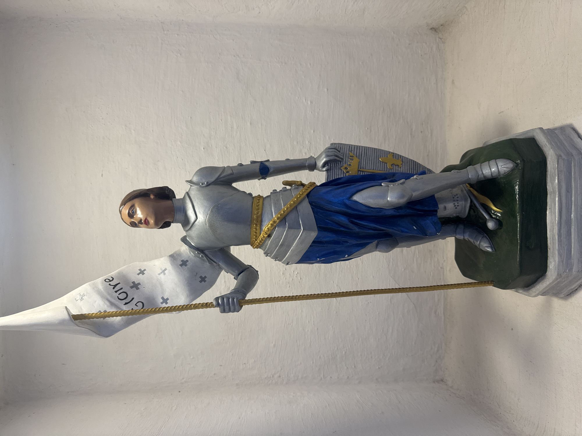 The image shows a statue of Joan of Arc, a historical figure. She is depicted as a warrior, wearing a suit of armor and holding a flag. The statue is standing on a base, which is on a shelf. The background is a white, textured wall. The statue is detailed, with realistic features. The flag has text and symbols on it. It is not possible to determine the location in Niterói, Brazil from the image.
