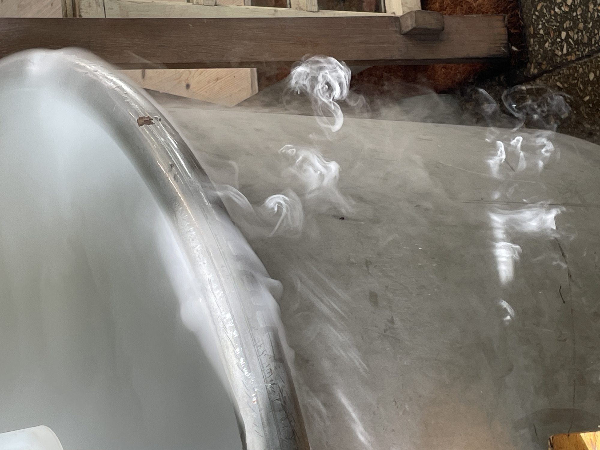 The image captures an indoor scene dominated by a large, light-colored, curved object, possibly a tank, pipe, or part of a machine. This object appears to be wrapped in a transparent plastic film, exhibiting wrinkles and reflections. A notable detail on its curved surface is a patch of brown-orange discoloration or damage.
White, dense smoke or vapor is a prominent feature, originating from or around the main object and spreading across the adjacent surfaces. The vapor forms distinct swirling plumes and tends to cling low to the ground and the object's surface, characteristic of cold vapor (e.g., from dry ice or liquid nitrogen).
In the background, wooden structures, including planks or beams, are visible, suggesting a workshop or industrial setting. Below the wooden elements, there's a dark, textured material that resembles bark mulch or similar organic debris. The main object rests on a light grey, somewhat reflective surface, likely concrete or a similar flooring.
No people or visible text are present in the image. The time of day and weather conditions are not discernible due to the indoor setting. While the location context is provided as Saint Petersburg, Russia, there are no specific visual cues within the image itself to confirm this. The overall impression is one of an experiment, a industrial process, or an unusual atmospheric effect.