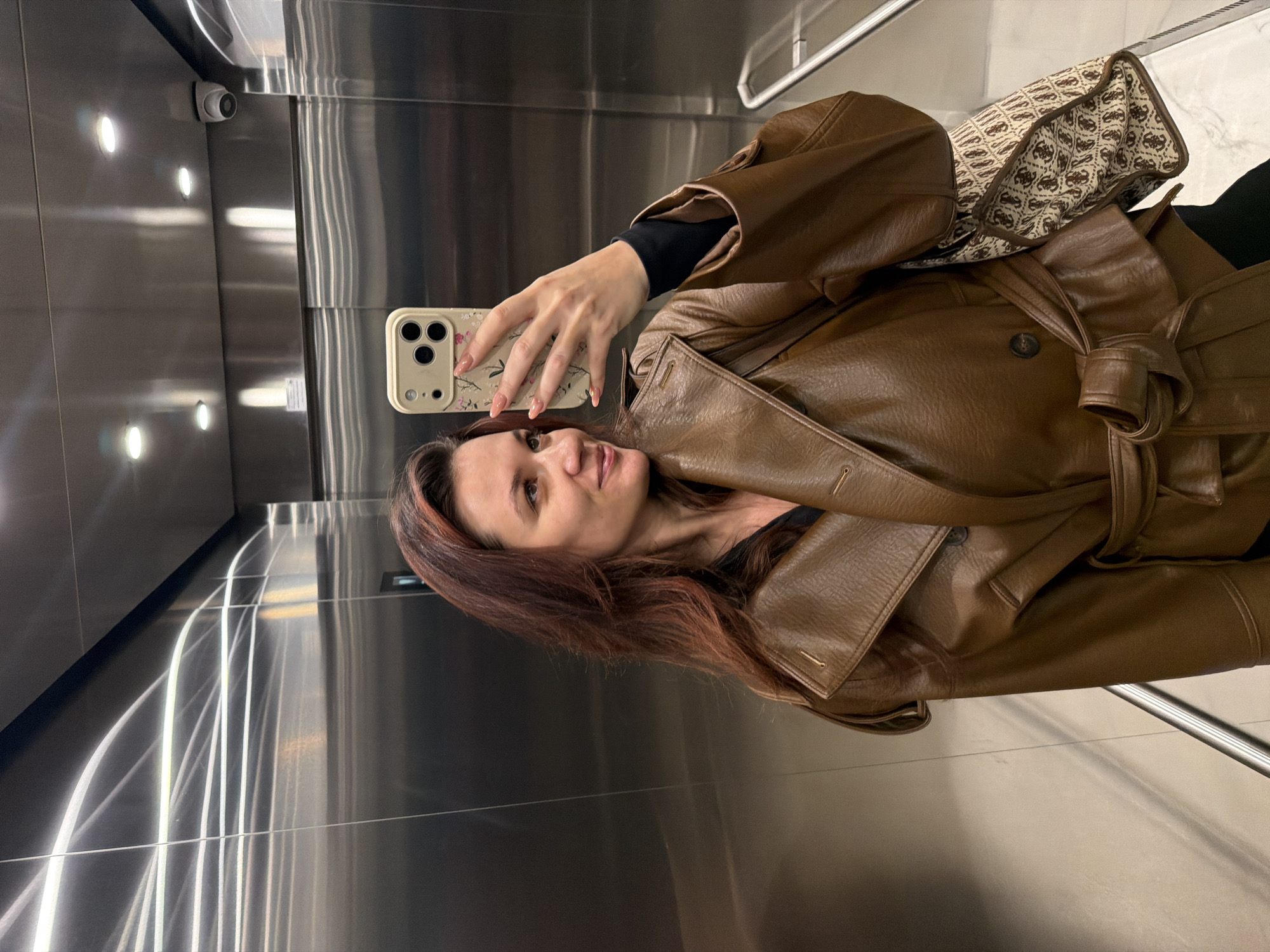 A woman stands in a modern elevator with stainless steel walls and recessed lighting. She is taking a selfie with her smartphone, which has a floral patterned case. She is wearing a brown leather trench coat, black undershirt, and carrying a handbag over her shoulder. The location appears to be Belgrade, Serbia.