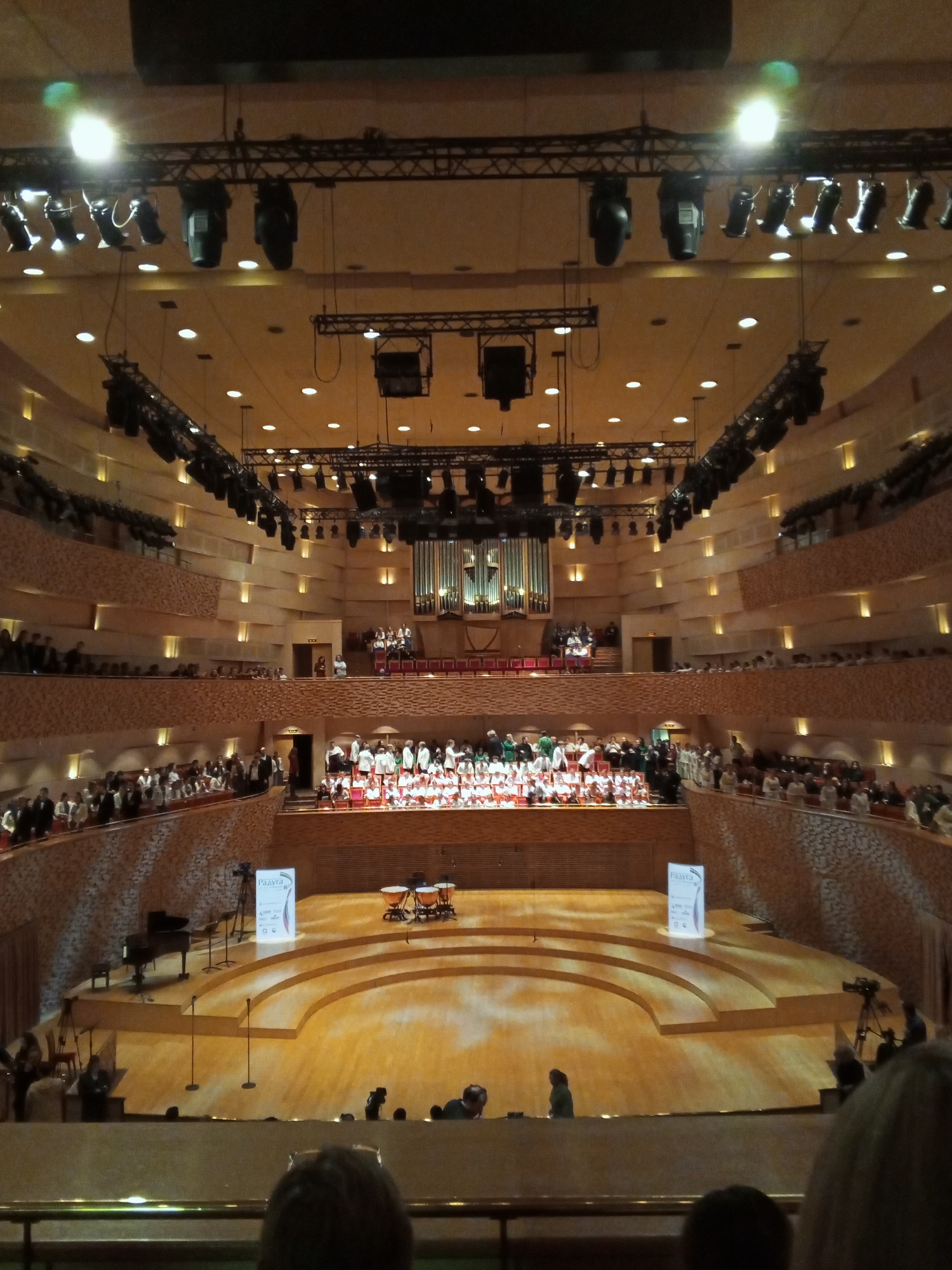 The image depicts a large concert hall, possibly in Saint Petersburg, Russia, based on the Cyrillic text on signage on stage. The hall is multi-tiered with audience seating in the balconies. A choir and other performers are assembled on the stage. An organ is visible in the background, behind and above the performers. Stage lighting rigs hang from the ceiling. A piano and timpani are on the stage. Several audience members are visible in the foreground.