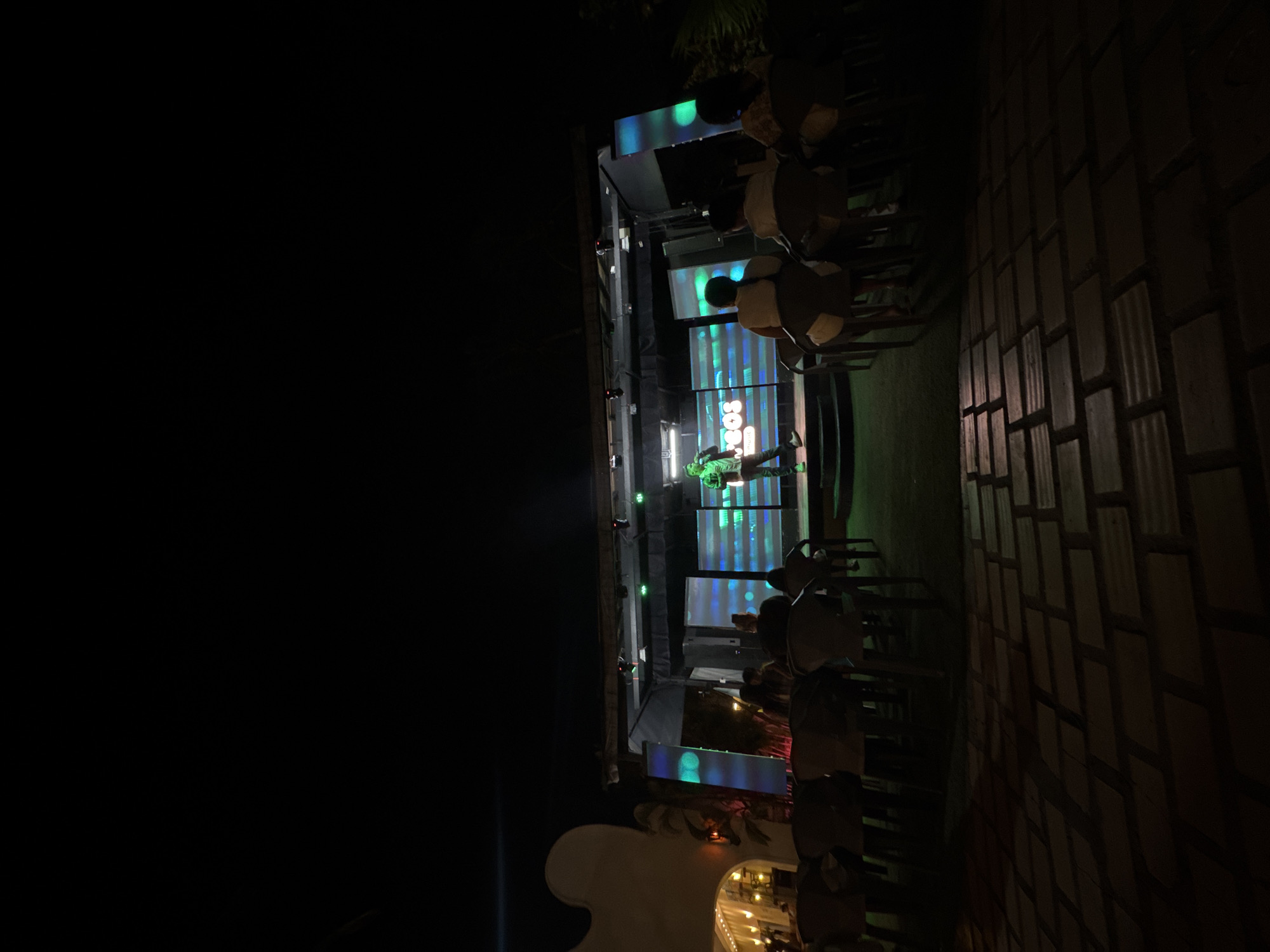The image depicts an outdoor performance stage at night, likely in Porlamar, Venezuela, based on architectural details. A musician is visible on stage, illuminated by green stage lights. A video screen with abstract visuals serves as a backdrop. An audience, seated on chairs, observes the performance. The surrounding environment is dimly lit, with brick or tile flooring and architectural elements suggestive of a resort or outdoor event space. The sky is dark, indicating nighttime.