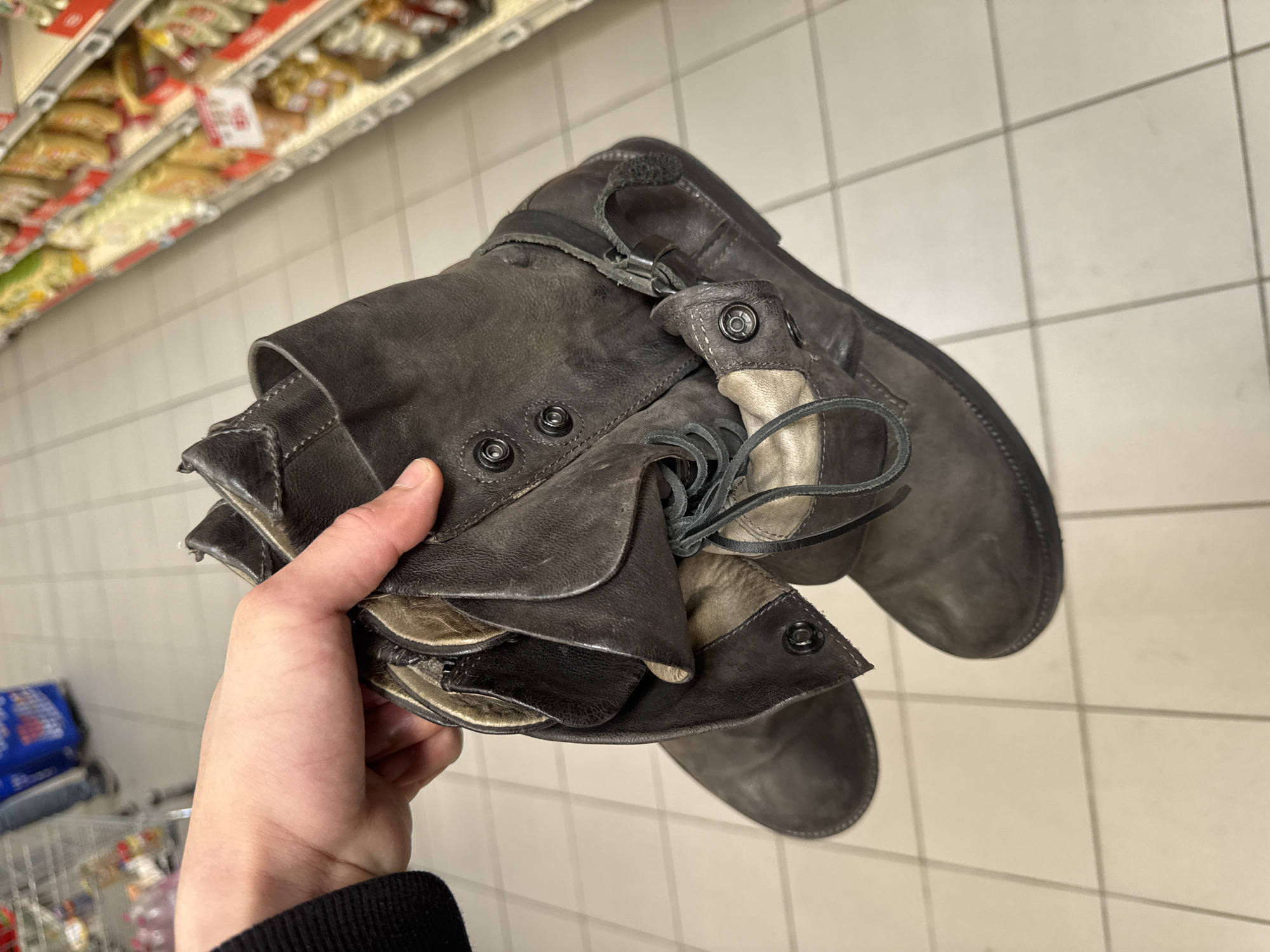 The image captures a close-up, high-angle view of a person's hand holding a pair of dark, distressed leather boots or shoes in an indoor commercial setting. The footwear is predominantly dark grey or dark brown, featuring a worn texture, laces, and multiple metallic snap fasteners and buckles on straps. A lighter, possibly beige or tan, material is visible as part of the interior lining or trim. The hand holding the boots has light skin, with a black sleeve visible at the wrist.
The background suggests a supermarket or large retail store, identifiable by the light-colored tiled floor with grey grout lines and out-of-focus shelves stocked with various products, some with unreadable red labels. A partial view of a shopping cart is visible in the lower-left corner. The scene takes place in Les Ulis, France. The activity appears to be the handling of merchandise, possibly during a shopping trip. Due to the indoor setting, the time of day and weather are not discernible. No legible text is visible within the image.