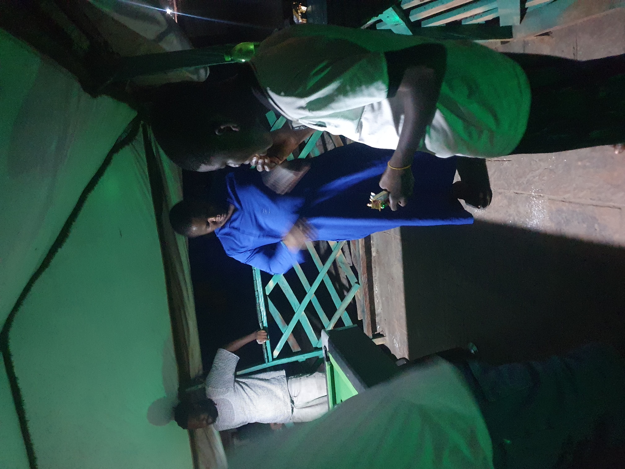 In Lokoja, Nigeria, a nighttime scene unfolds under a green awning. Three men are present. On the left, a man with dark hair and a beard wears a checkered short-sleeved shirt and light-colored shorts. He stands near a green pool table. In the center, another man, also with dark hair, is dressed in a vibrant blue robe. He appears to be in motion. To the right, a third man with dark skin and hair wears a light-colored t-shirt and dark pants. He is holding a small object in his right hand and appears to be eating something. A green railing is visible behind the central and right figures, and beyond that, a dark background with some distant lights. The ground is paved and appears somewhat wet. The overall lighting is dim, with a distinct green hue cast by the awning overhead.