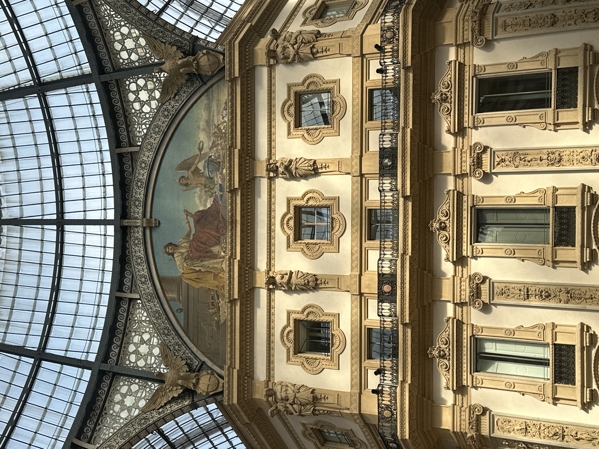The image captures a section of the Galleria Vittorio Emanuele II, a historic shopping arcade located in Milan, Italy. The architecture is ornate and features a prominent arched fresco at the top, depicting allegorical figures. Below the fresco, the building's facade is adorned with decorative sculptures of eagles with spread wings flanking the fresco, and other human figures integrated into the architectural elements. The facade also displays several large windows with decorative framing and balconies with intricate iron railings. The upper part of the image shows the glass and metal structure of the Galleria's dome, allowing natural light to filter through. The building material appears to be light-colored stone or stucco, with golden accents highlighting the elaborate details. The scene is static and focuses solely on the architectural grandeur.