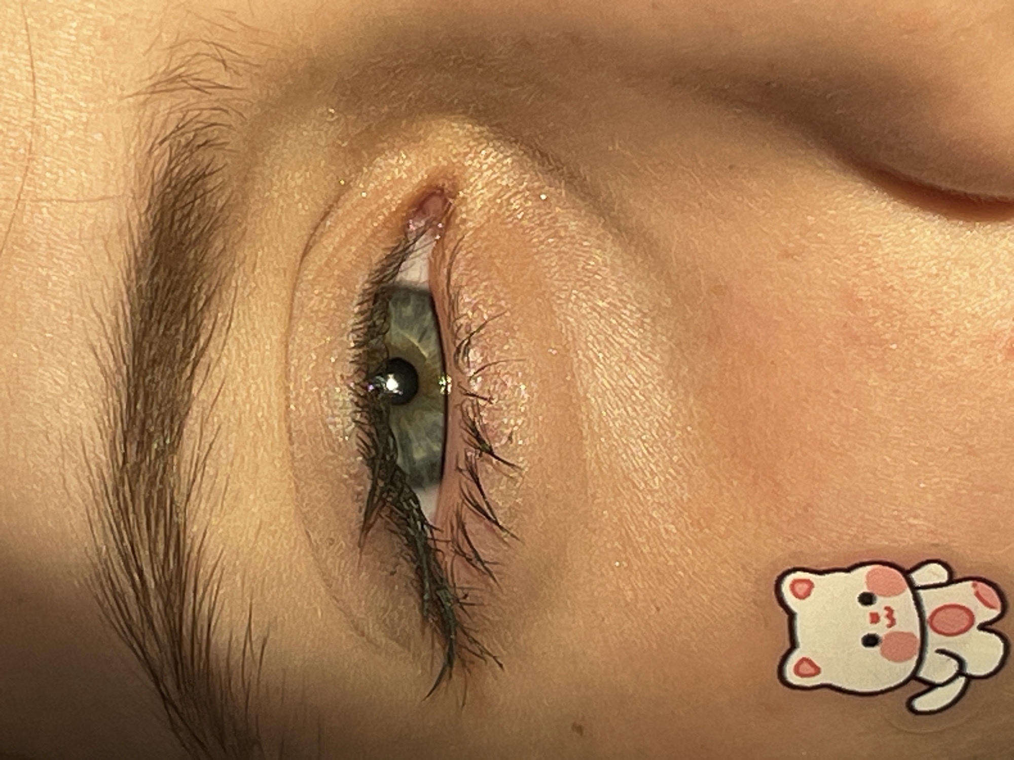 The image shows a close-up of a person's closed eye, brow, and adjacent skin. The person has a light skin tone, and visible eyebrow hairs. The eye has eyelashes and faint light green irises. Light reflects off the eye, creating shiny spots. There's also a small teddy bear sticker on the skin near the eye. No information in the provided image confirms a location in Izhevsk, Russia.