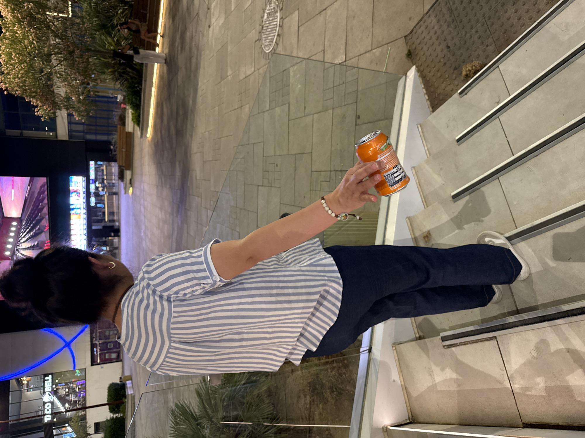 A person is standing on a set of outdoor stairs at night, holding an orange soda can. The person is wearing a blue and white striped shirt and dark pants. They have dark hair tied back and are wearing a small gold hoop earring. The background shows an outdoor area with a glass railing, tiled flooring, and some trees. In the distance, there are illuminated signs for businesses, including "TACO BELL". The scene suggests an urban or commercial setting. The lighting indicates it is nighttime.