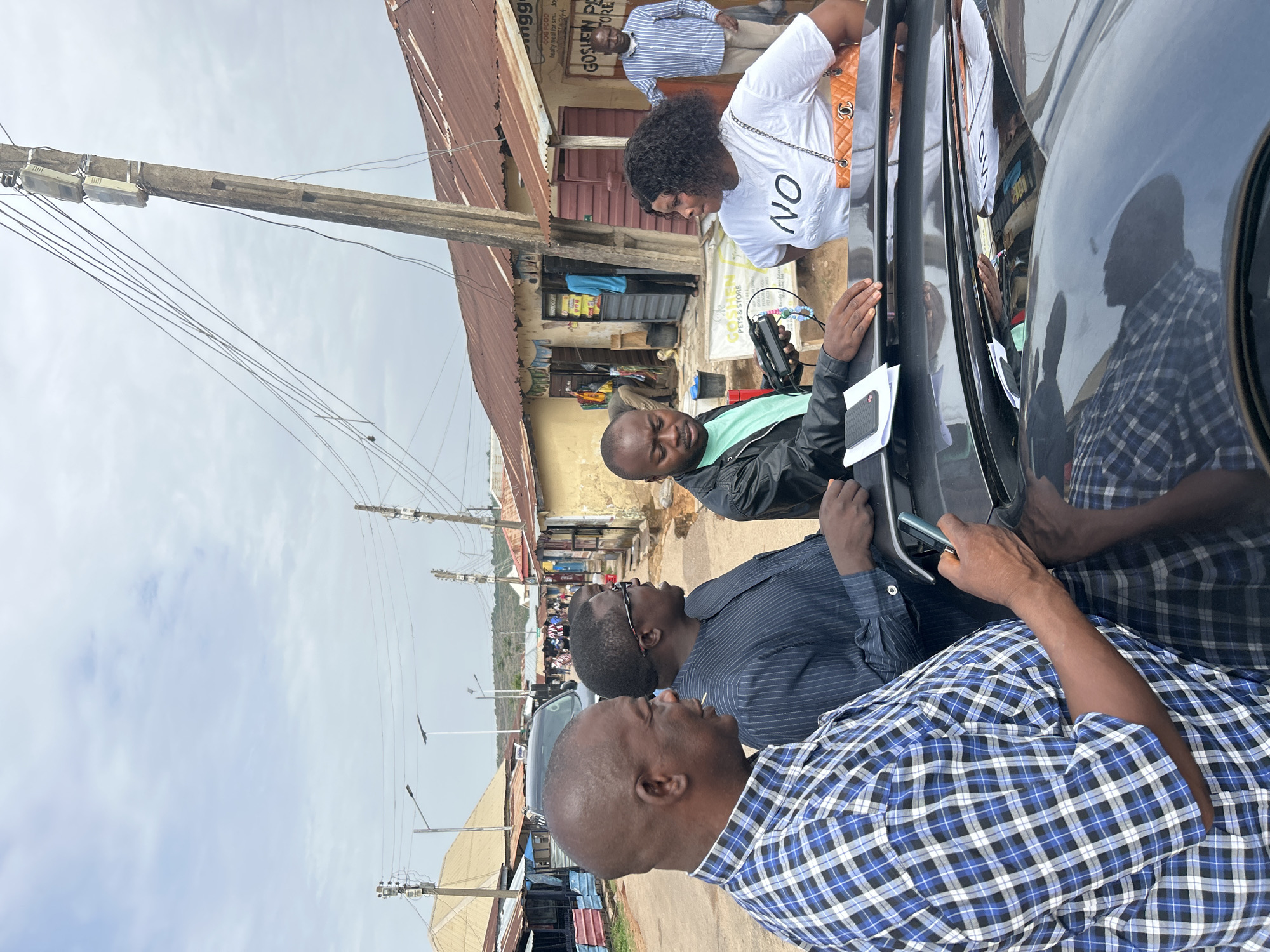 The image shows a group of four people standing near a dark-colored vehicle on a street in Jos, Nigeria. A woman in a white shirt is leaning toward the vehicle, and a man in a black jacket is holding a device near the vehicle. Two men in button-down shirts are standing and watching. Behind them are buildings with signs for "Goshen Pets & Store" and other businesses. Utility poles with wires are visible overhead. Other vehicles are parked along the street.
