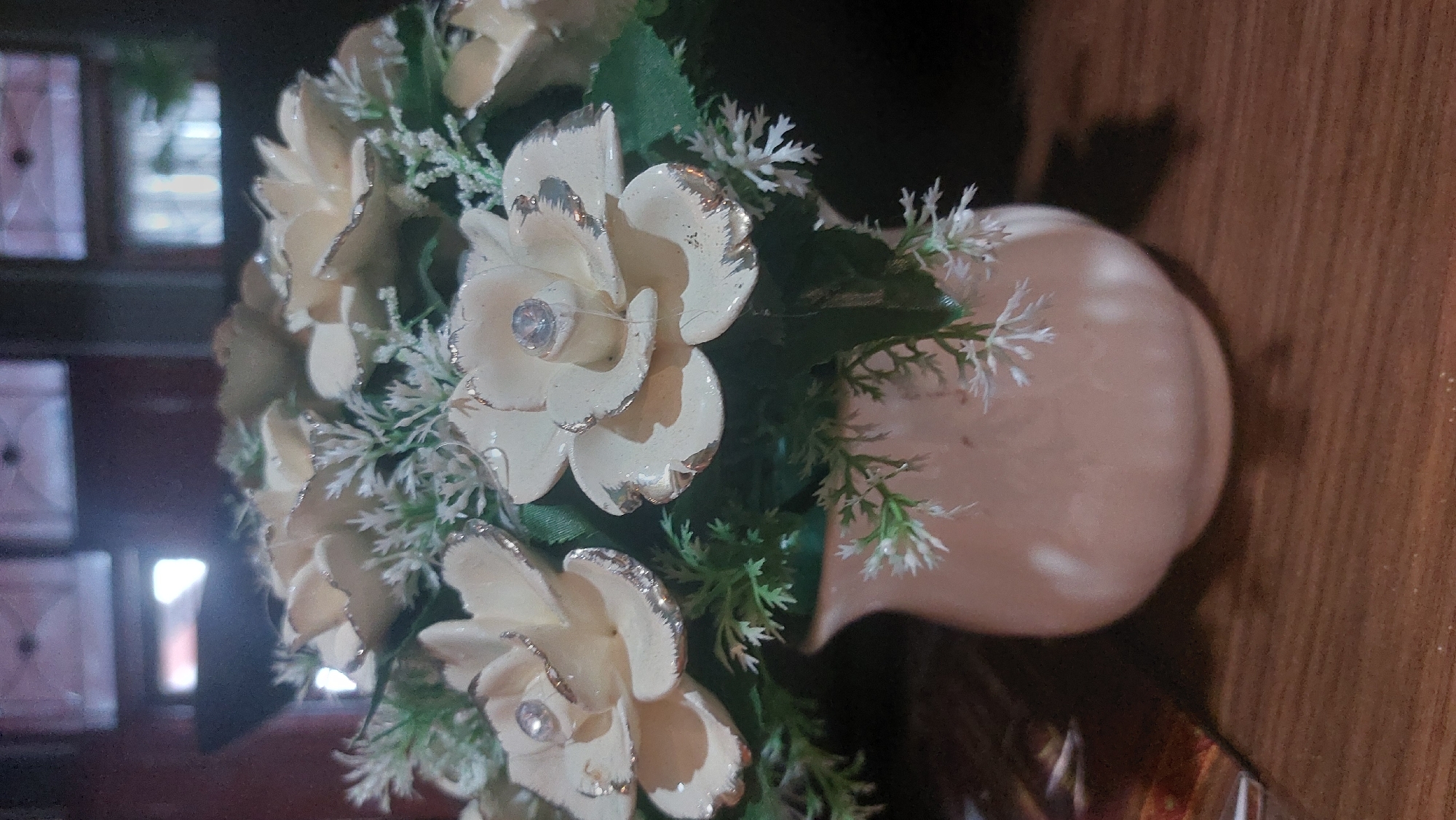 An indoor, close-up shot captures a decorative floral arrangement in a light pink ceramic vase. The arrangement features several cream-colored artificial roses, each adorned with a small, clear rhinestone in the center and silver detailing on the edges of its petals. Delicate white artificial filler flowers and green artificial leaves fill the spaces between the roses, creating a lush appearance. The vase has a smooth, matte finish and a bulbous shape that tapers to a narrower opening. The arrangement rests on a wooden surface, likely a table, which displays a visible wood grain pattern. In the blurred background, a dark, out-of-focus area suggests an indoor setting, possibly a room with windows or framed elements, though specific details are indistinct. The lighting is soft, with some light reflecting off the vase and the rhinestones on the roses. The image does not contain any people or specific landmarks that would allow for definitive location identification within the city of Medan, Indonesia.