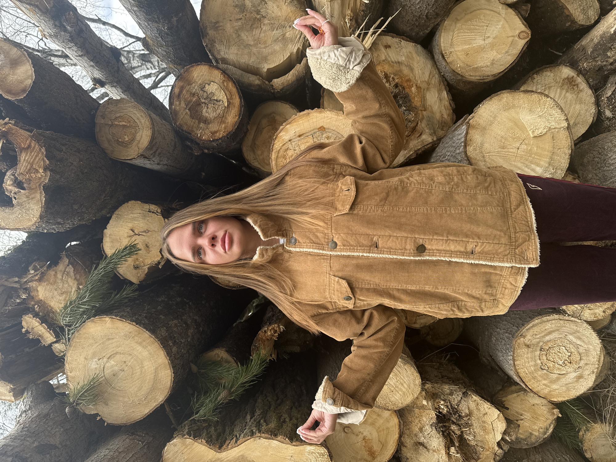 A young woman lies among a pile of logs. She has long blonde hair and is wearing a brown corduroy jacket with a sherpa lining. She is also wearing dark purple pants. Her right arm is raised. The background is a pile of logs and tree branches. The location is possibly Grebnevo, Russia.