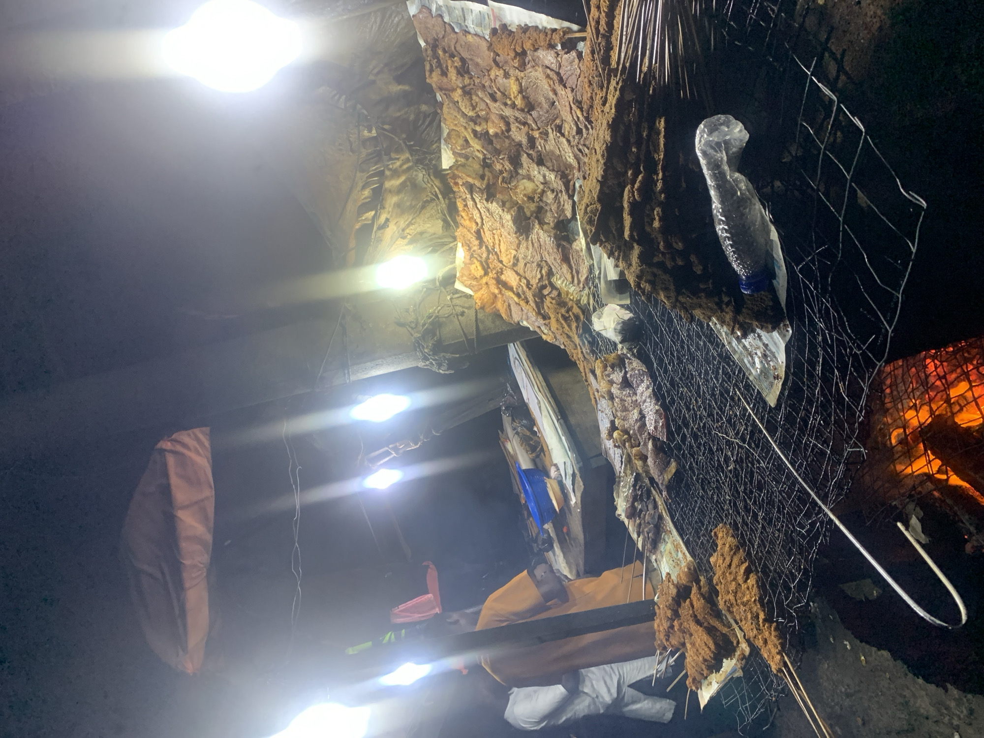 The image depicts a nighttime scene at a street food stall, likely in Port Harcourt, Nigeria, with food being grilled over an open fire. The stall is illuminated by several bright, overhead lights, casting harsh shadows and highlighting the food and the surrounding environment.
In the foreground, a large metal grill is set over glowing embers, with various skewers of seasoned meat being cooked. The meat appears to be in small, bite-sized pieces, some coated in a dark spice rub. Further back, a large, flat surface is covered with thinly sliced, dried meat that is also being grilled or warmed. The meat on this surface is a reddish-brown color and appears to be quite dry.
To the left, two individuals are partially visible. One is wearing a white garment, and the other is wearing an orange t-shirt. They appear to be interacting with the food or the stall owner.
The stall itself is covered by a makeshift roof made of fabric or plastic sheeting, which sags in places and is supported by wooden structures. There are various containers and supplies scattered around, including a blue bowl on a wooden counter. A partially empty plastic water bottle is visible on the grill in the mid-ground. The overall impression is one of a busy, open-air food market at night.