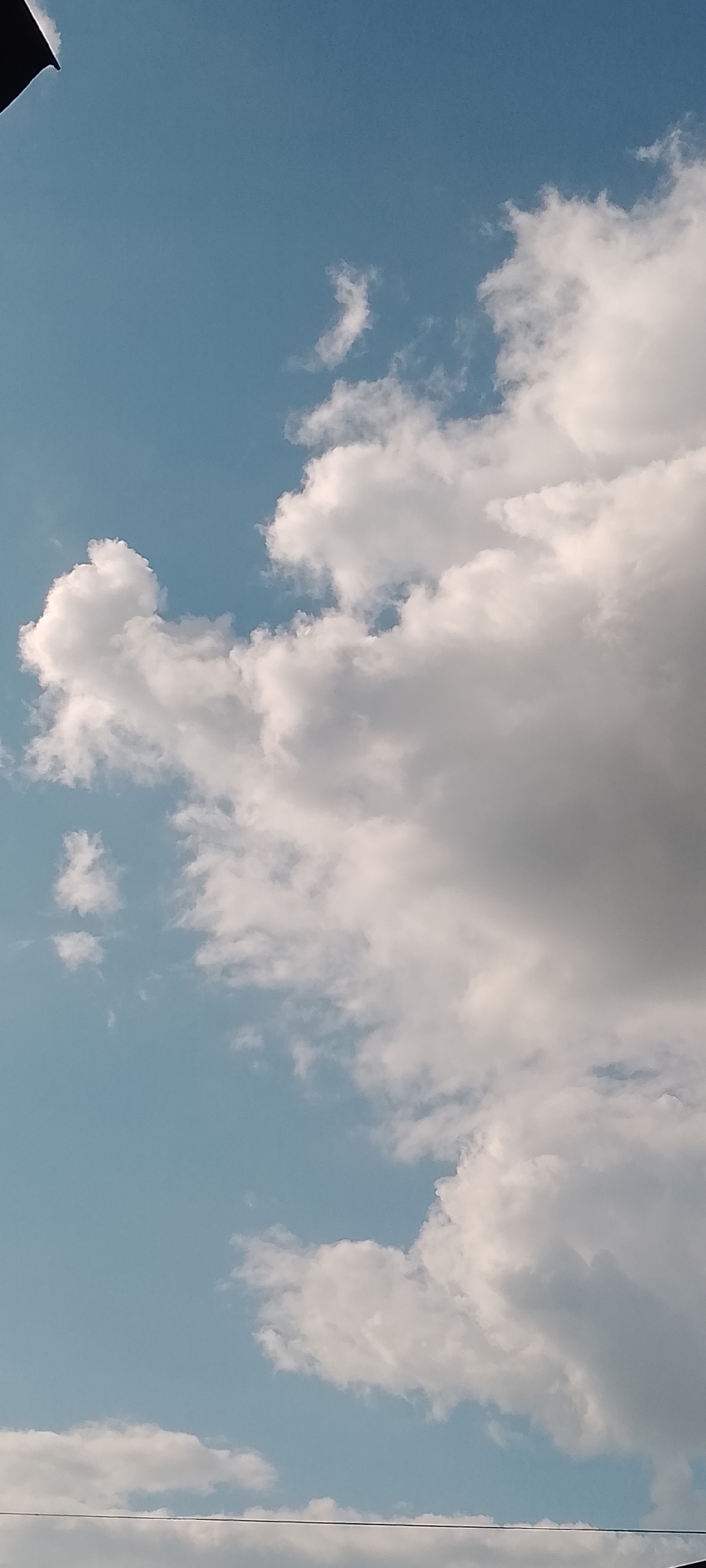 The image shows a close-up view of a clear blue sky with fluffy white clouds. There are no people or identifiable objects in the frame besides the clouds and the sky. The lighting suggests it is daytime, and the angle of the shot is from below, looking upwards. There are also faint horizontal lines visible at the bottom of the image, which might be power lines or wires. The scene is generic and does not provide any specific information about its location in a city or otherwise.