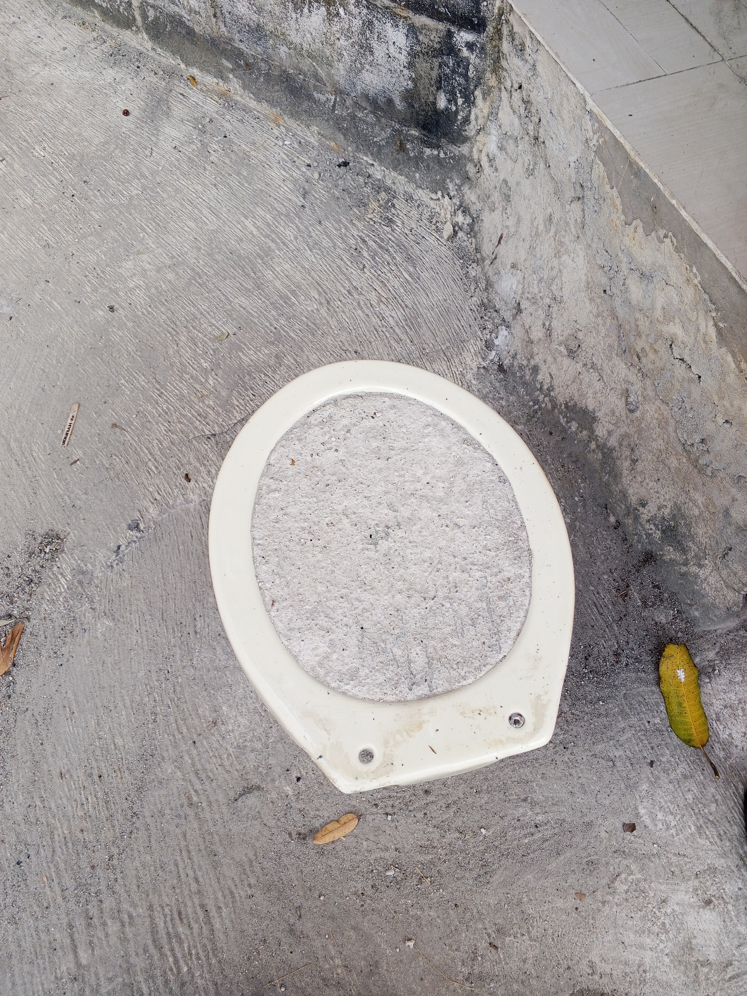 The image displays a white toilet seat with a concrete interior, resting on a rough, textured concrete surface. The surrounding environment appears to be an outdoor area, possibly a public space or under construction, given the exposed concrete and scattered debris. Several dried leaves are visible on the ground, along with some dark specks that could be dirt or small stones. In the upper right corner, a portion of a textured wall or structure is visible, with signs of wear and discoloration. A single yellow leaf with a visible vein pattern lies to the right of the toilet seat. The overall scene is static and devoid of human presence. The location is identified as Ciruas, Indonesia.