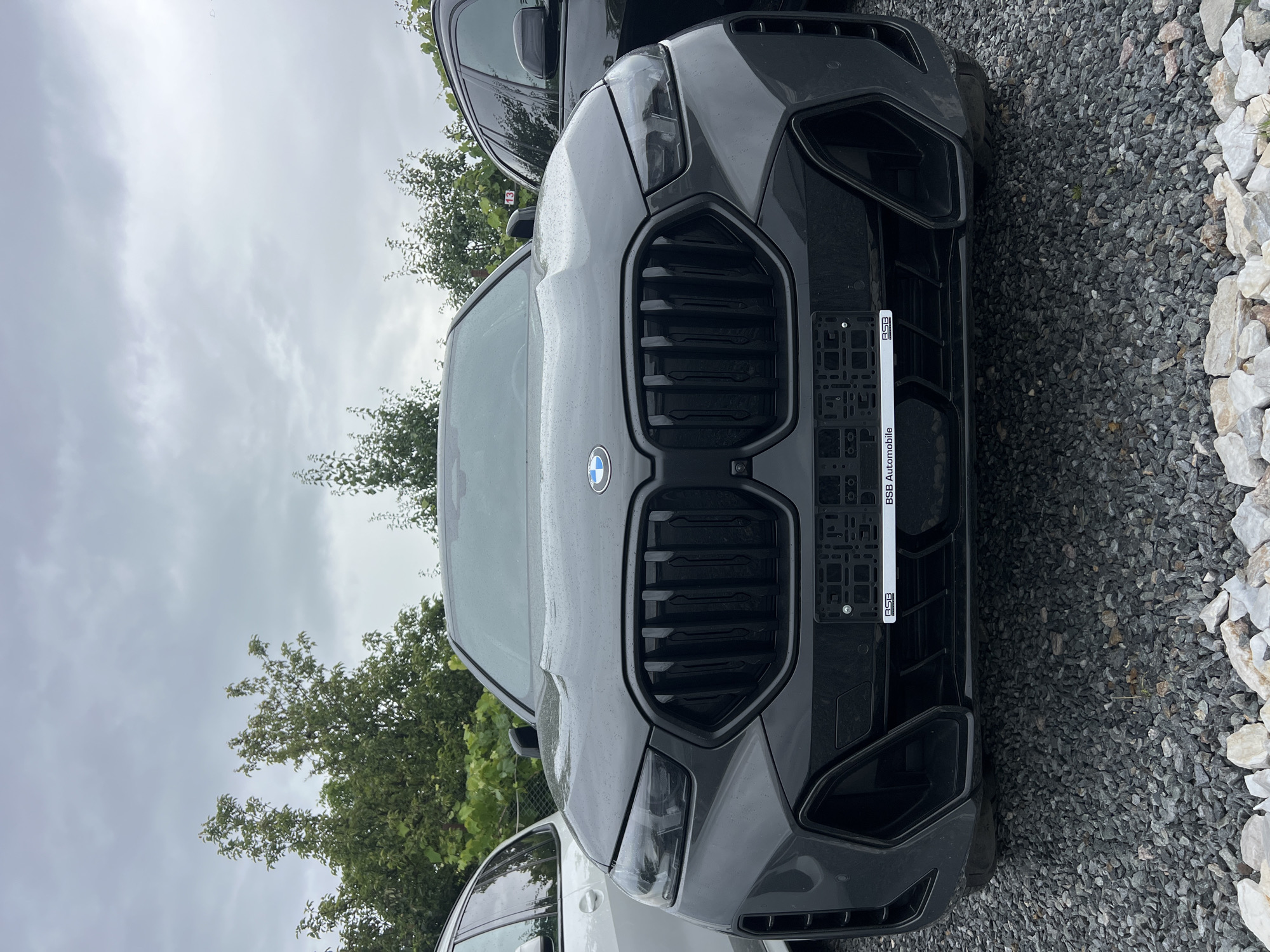 A front-facing, eye-level shot captures the grille and headlights of a dark grey BMW SUV parked on a gravel lot under an overcast sky. The car's large, vertical grille dominates the center of the frame, with sharp, angular headlights flanking it. A license plate holder with the text "BSB Automobile" is mounted centrally below the grille. To the left, a portion of a white car is visible, and to the right, the dark body of another vehicle is partially obscured. Lush green trees form the background. The scene appears to be an outdoor car lot or dealership.