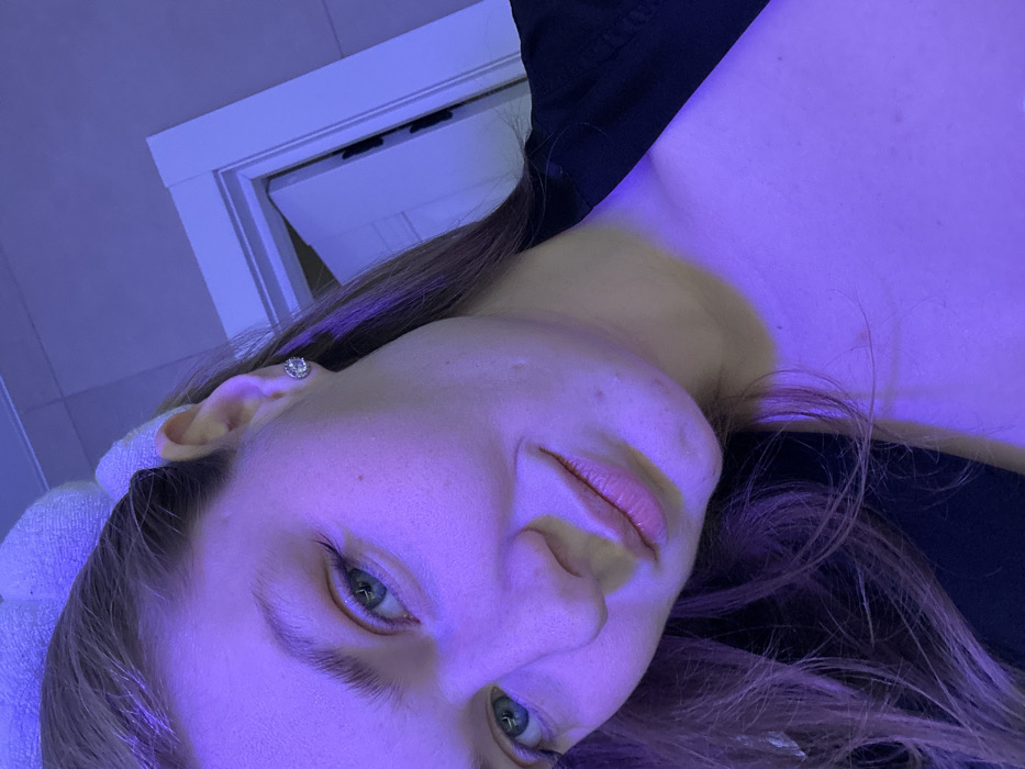 The image is a close-up portrait of a person, likely female, captured with a significant tilt, presenting the subject as if lying down with their head on the left side of the frame and their upper body extending towards the right. The entire scene is bathed in a distinct purple-blue ambient light, creating a cool and somewhat ethereal atmosphere.
The subject has fair skin, which is strongly colored by the purple light, and notable green or hazel eyes that are open and looking directly at the viewer. Reflections of the blue light are visible in the irises. Long, dark brown or auburn hair frames the face and falls across the neck and shoulder area. The left ear is visible, adorned with a silver-colored stud earring that features a central clear gemstone surrounded by smaller, sparkling stones. The person's lips are slightly parted, revealing a natural pink hue, contrasting subtly with the purple illumination on the skin. A dark-colored garment, possibly black, is visible on the upper body and shoulder.
In the background, a small portion of what appears to be a white door frame or wall trim is visible towards the top-left corner of the image, suggesting an indoor setting. The rest of the background is indistinct and obscured by the strong purple lighting. The image focuses primarily on the person's face and upper chest. There is no specific activity taking place other than posing for the photograph, and no visible text is present in the image. The lighting choice gives the portrait a stylized and moody aesthetic.