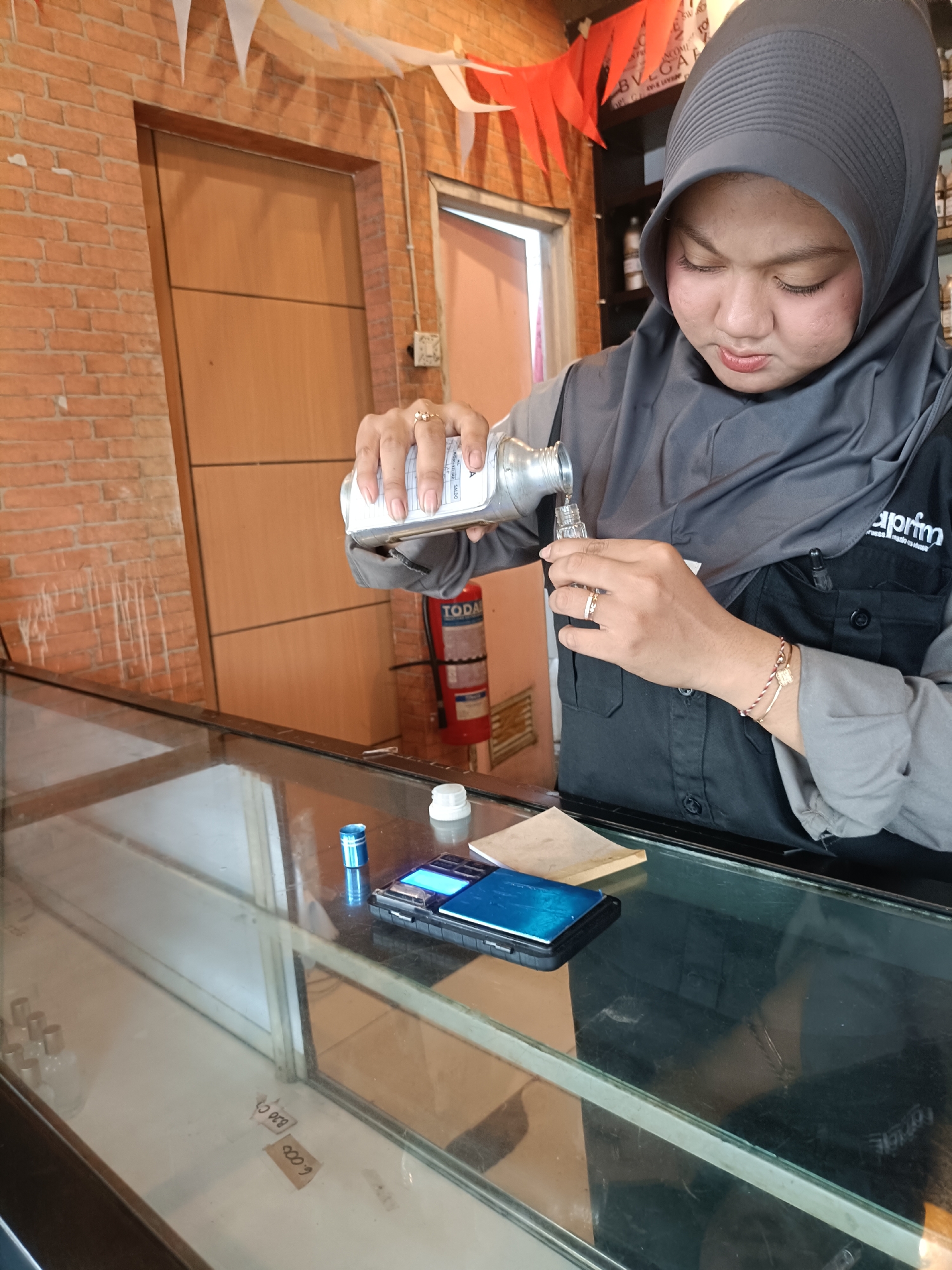 The image shows a woman wearing a grey hijab and a black uniform shirt, pouring liquid from a metal bottle into a small glass bottle. She is standing behind a glass counter. On the counter, there are a few items including a mobile phone, a small blue cylindrical object, and a white cap. In the background, there is a brick wall, a wooden door, and a red fire extinguisher. The scene appears to be set in a retail or workshop environment. The text on the woman's shirt is partially visible and seems to say "aprim". The overall setting is indoor, possibly a shop in Kediri City, Indonesia, judging by the possible Indonesian context suggested by the presence of the brand and the implied cultural attire.