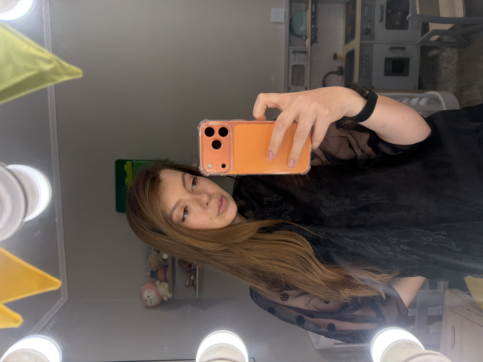 A woman takes a selfie in front of a vanity mirror with lights. She is holding an orange phone in her right hand and is wearing a black sheer top with polka dots. Her hair is long and brown. The background shows a partially visible playroom with a toy kitchen set and other toys. The location is Unknown, Unknown.