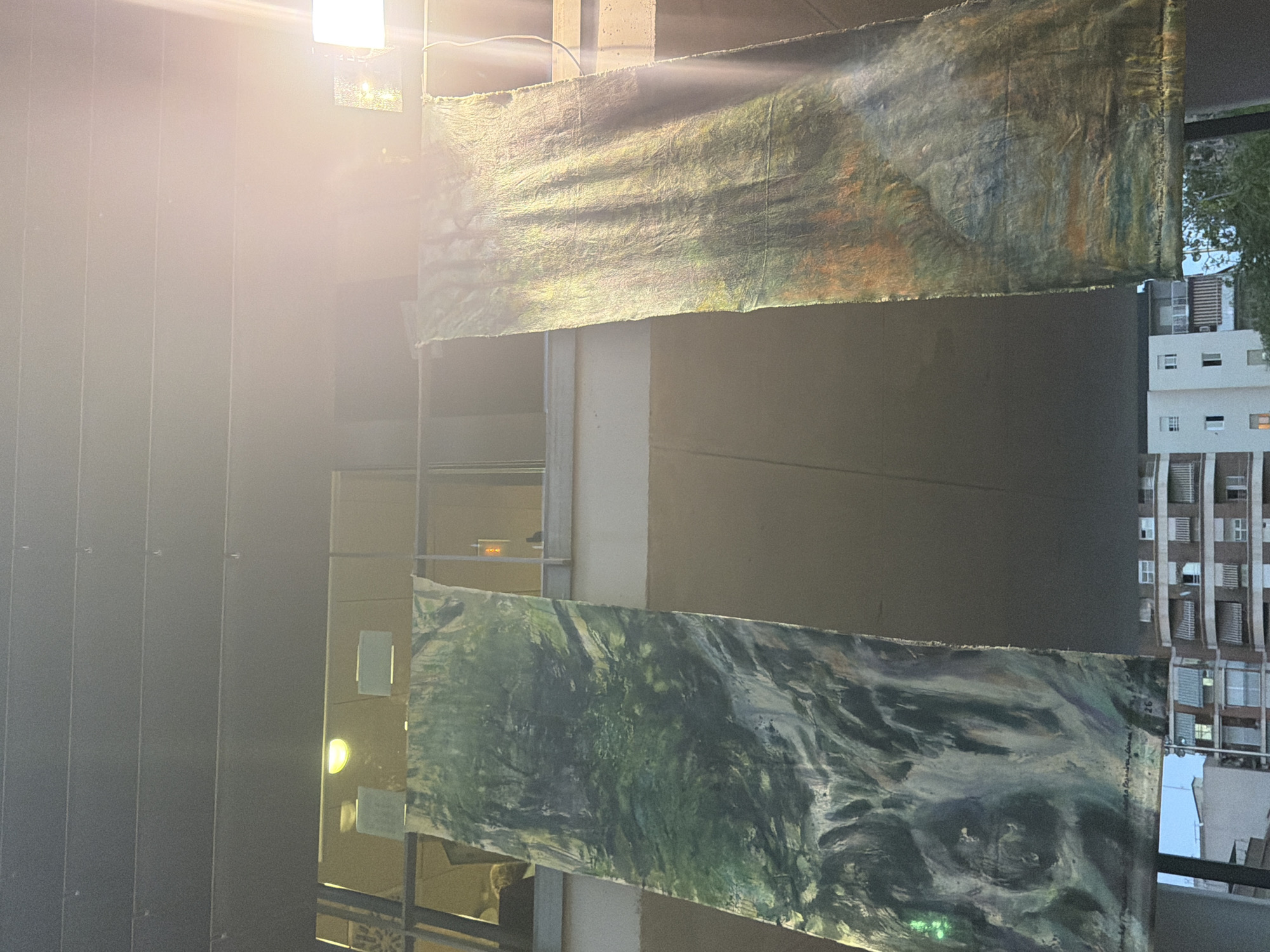 The image shows two large, rectangular tapestries hanging vertically. The tapestry on the left depicts a dark green, abstract landscape with swirling brushstrokes that suggest trees and water. The tapestry on the right features a more colorful, abstract composition with shades of green, orange, and yellow, also appearing to be a landscape. Both tapestries have a distressed, textured appearance. They are suspended from a horizontal bar, which is part of a building's exterior structure, possibly a balcony or overhang. Behind the tapestries, a windowed building facade is visible, with interior lights suggesting an indoor space. The background shows the sky and the upper floors of other buildings. A bright light source is visible in the upper right corner, causing lens flare and overexposure. The scene appears to be outdoors, in an urban environment, possibly a public space or an art exhibition. The text on the bottom of the left tapestry appears to be a signature and date, with visible words "Mundo" and "2026". The text on the bottom of the right tapestry is also a signature and date. Given the context of the provided image, this scene is located in the city of Elche, Spain.