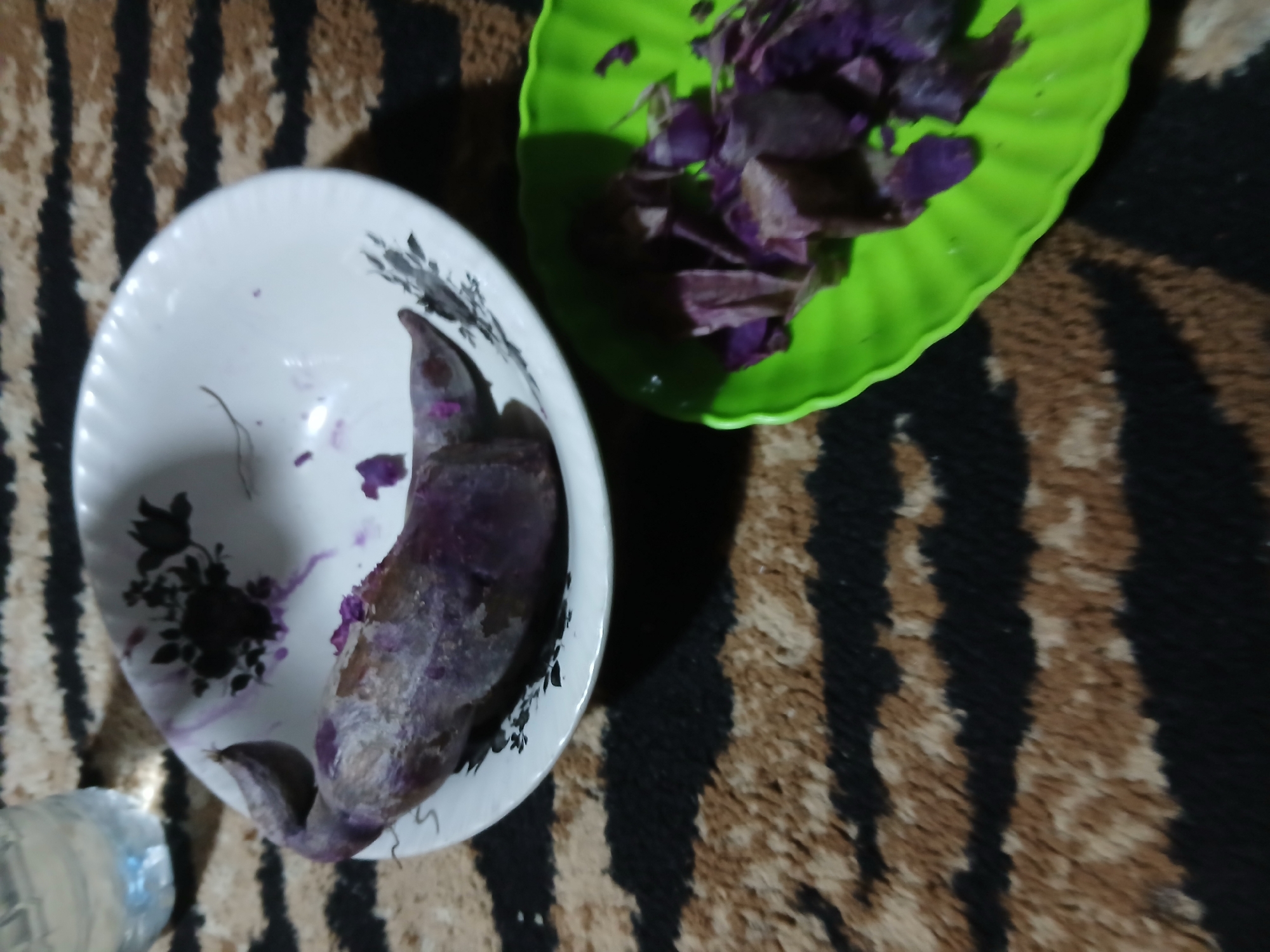 The image shows a close-up of two plates of purple sweet potato on a striped rug. One plate, a white bowl with a floral design, contains a whole purple sweet potato. The other plate, a green wavy dish, contains cut pieces of purple sweet potato. A partially visible glass of water is in the background. The scene appears to be indoors. The location in the city of Demangan, Indonesia is not discernible from the image itself.