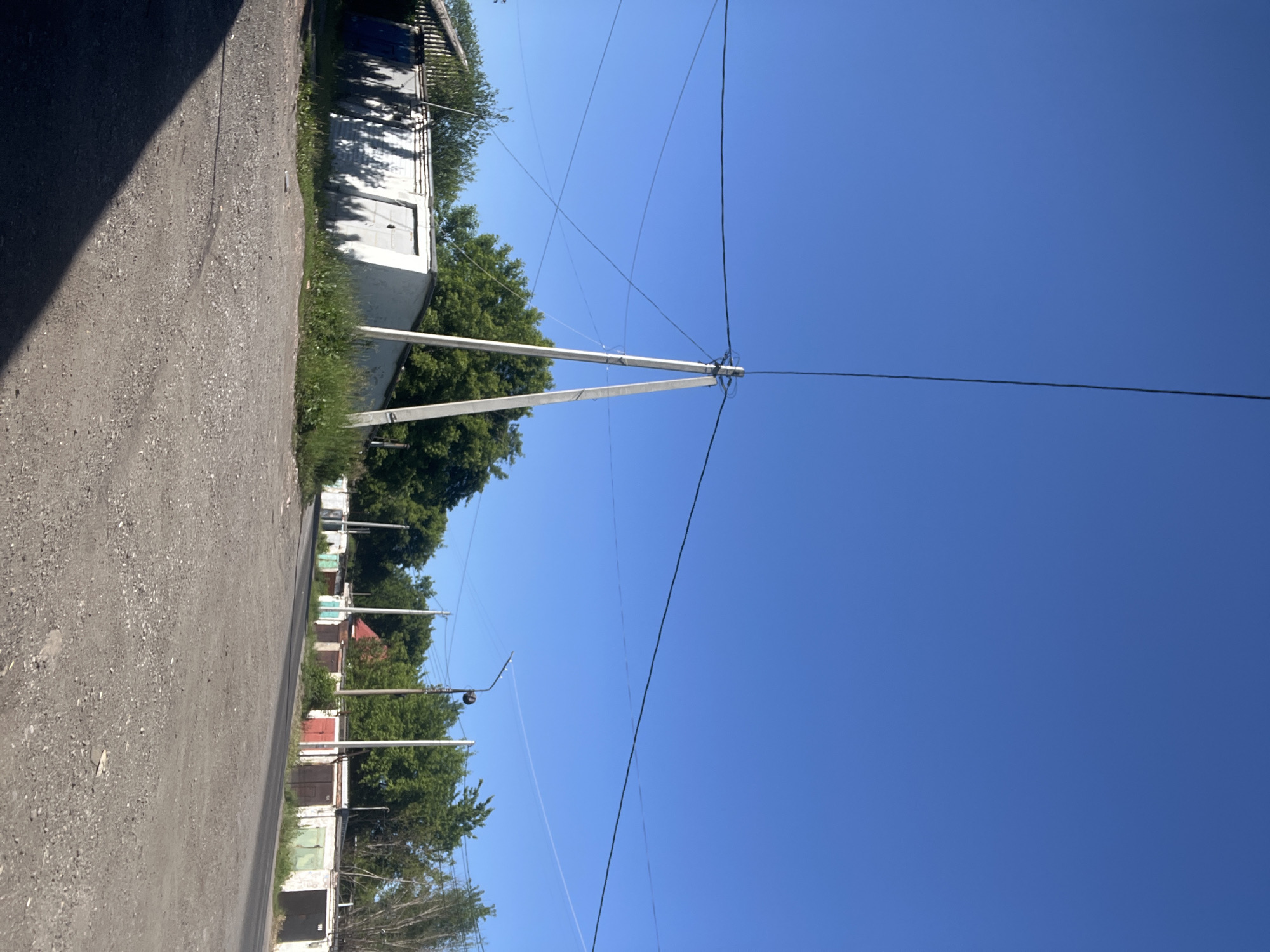 This media file depicts an outdoor scene in Kiselyovsk, Russia, under a clear, bright blue sky, suggesting it is daytime with good weather. The perspective is low and angled, looking up towards the sky and along a gravel road.
The scene is characterized by a row of garages lining one side of the road. These garages appear to be utilitarian structures, with varying colors and door types visible, some with white facades and others painted in muted tones of green, red, and brown. Overgrown green foliage and trees are visible behind and between the garages, indicating a somewhat unkempt or naturalized environment.
Dominating the upper portion of the image and extending across the sky are numerous overhead power lines and utility poles. One prominent pole, made of concrete or a similar material, is angled and supports multiple wires. The power lines crisscross the sky, creating a visually complex network.
The road itself is a rough, gravel surface, with a dark shadow cast from the left side of the frame, possibly from a building or a large tree not fully in view. The overall impression is that of a residential or industrial fringe area, likely a service road or access path to the garages. There is no visible text in the image. The scene evokes a sense of everyday life in a Russian town, with an emphasis on functional infrastructure and natural elements.