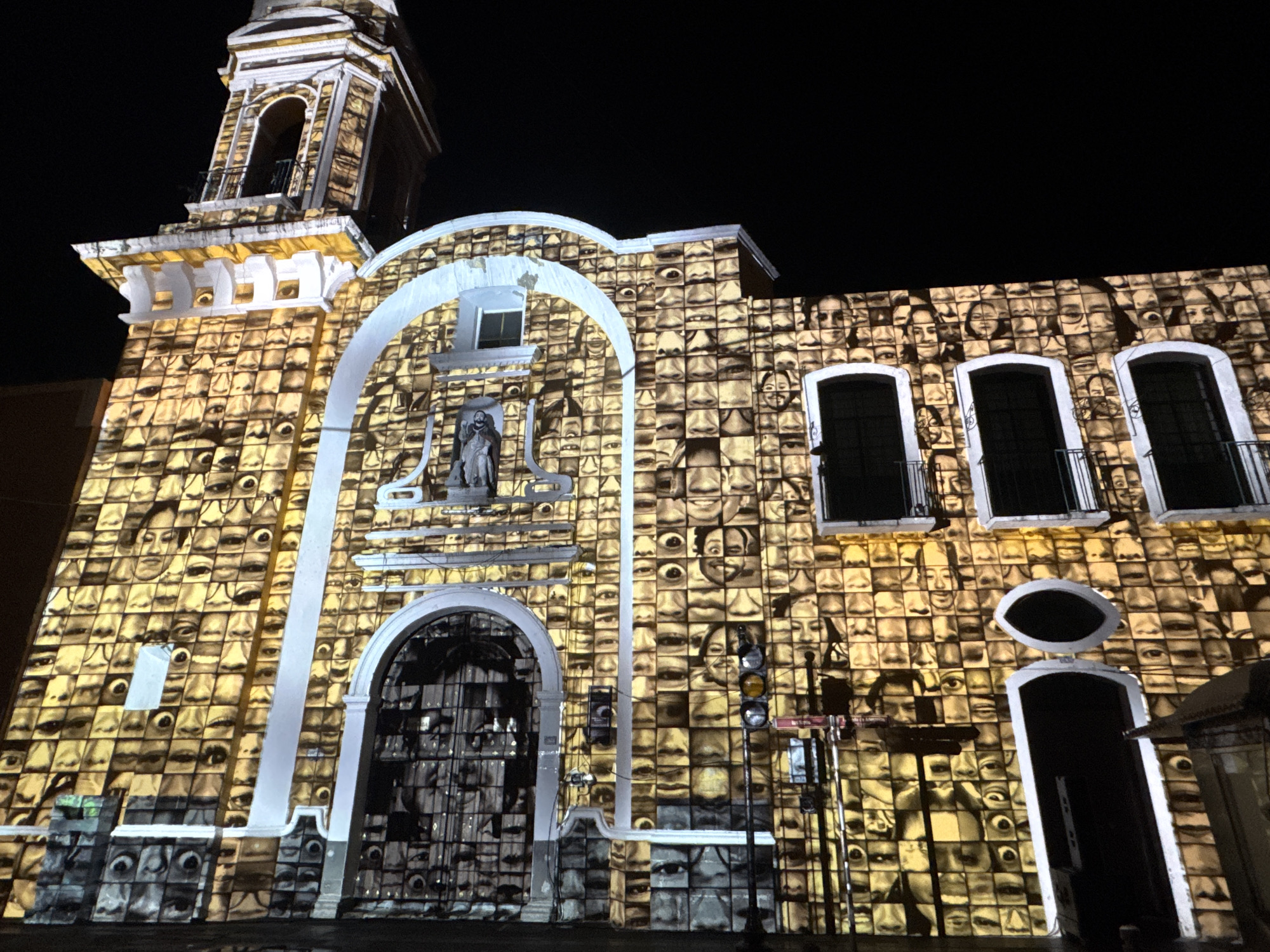 The image depicts the façade of a church in Puebla, Mexico, illuminated with a projection featuring numerous faces in a grid pattern. The church features a bell tower, an arched entrance, and several windows. A statue is visible above the main entrance. Traffic lights and street signs are present in front of the building, indicating an urban setting. The lighting projection gives the illusion that each brick of the church is a tile with a face on it.