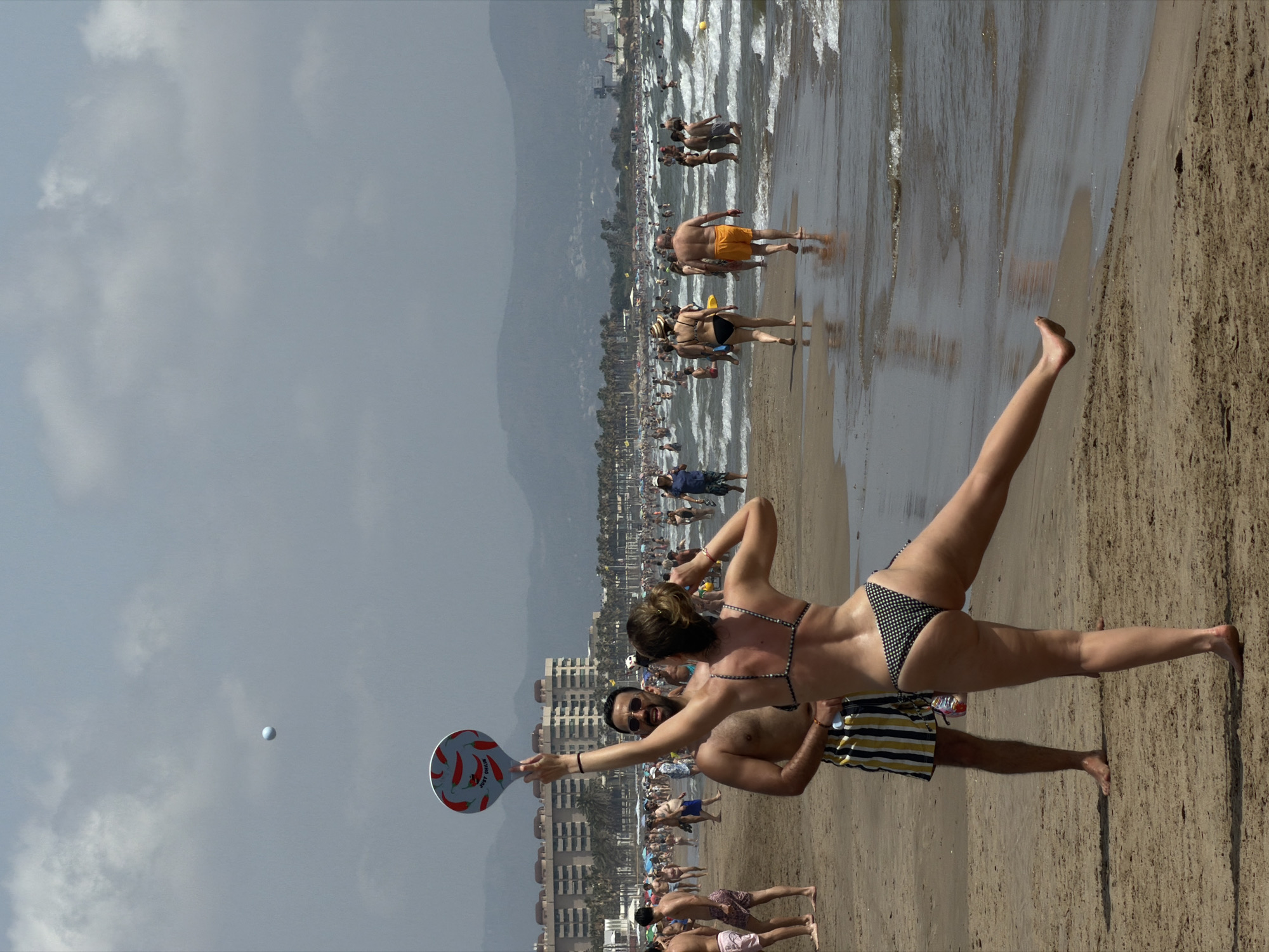 A group of people are on a crowded beach on a sunny day. In the foreground, a man and a woman are playing paddleball. The woman, in a black and white polka dot bikini, is in mid-air, reaching for the ball with her paddle. The man, wearing striped swim trunks, stands behind her. In the background, many people are walking along the shore, swimming in the ocean, and lounging on the sand. The ocean is choppy with small waves. In the distance, there are buildings and mountains. The sky is overcast with scattered clouds.