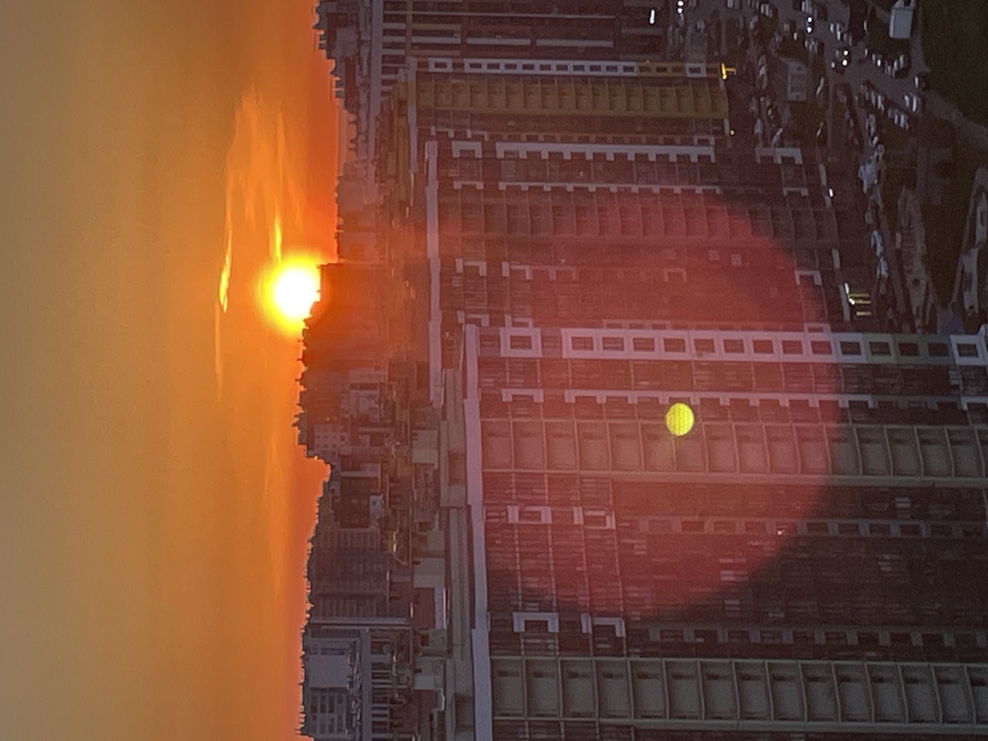 The image captures a striking sunrise or sunset over a bustling urban landscape, set in Saint Petersburg, Russia. The sky, dominating the left side of the frame, is awash in vibrant shades of orange and yellow, with the intense sun appearing as a brilliant, glowing orb positioned low on the horizon. The atmospheric conditions create a soft, ethereal haze that diffuses the sunlight, painting the entire sky in warm, fiery tones.
On the right, a dense collection of multi-story buildings, likely residential or commercial complexes, forms a jagged skyline. These structures feature repetitive patterns of windows and balcony-like elements, and their facades reflect the dramatic hues of the sky, appearing in shades of red, orange, and muted brown, with some sections highlighted by yellow architectural details.
A prominent lens flare is visible on the right side of the image, manifesting as a large, reddish-pink circular glow with a distinct smaller yellow-green circle superimposed over the buildings. In the upper right portion, a street is visible, populated with numerous cars, some displaying illuminated headlights or taillights, further suggesting the dawn or dusk hour. The scene vividly portrays the interplay of natural light and urban architecture at a significant time of day. No visible text is present in the image.
