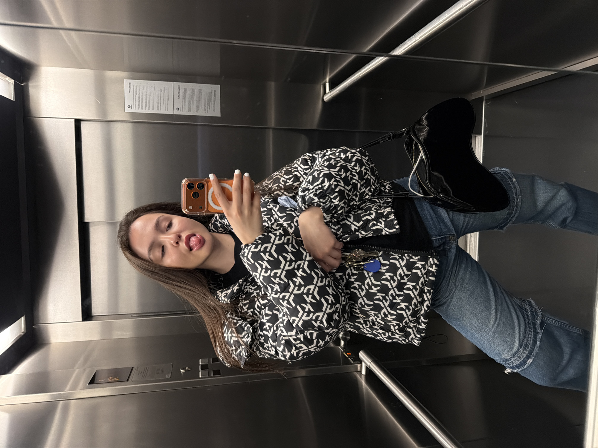 A young woman takes a selfie in a metallic elevator. She is wearing a black and white patterned DKNY jacket, blue jeans, and black boots with a matching black shoulder bag. She is holding an iPhone with a clear case. She is sticking her tongue out. The elevator has metallic walls and a control panel. The scene is in an elevator, but the location in Sofia, Bulgaria is unknown.