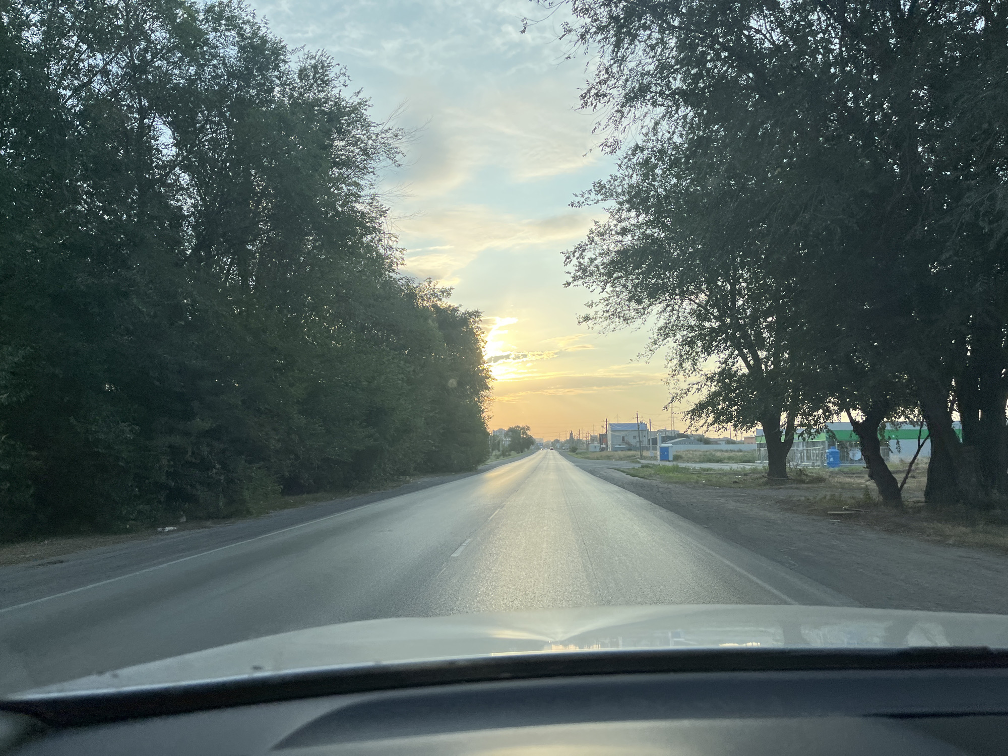 The image depicts a view from inside a vehicle driving on an asphalt road towards a horizon illuminated by the setting sun. Tall trees line both sides of the road, creating a canopy effect. In the distance, a few buildings and what appear to be utility poles are visible. The sky is partly cloudy, with streaks of orange and yellow light from the sun breaking through. The overall impression is that of a rural or suburban road at dusk.