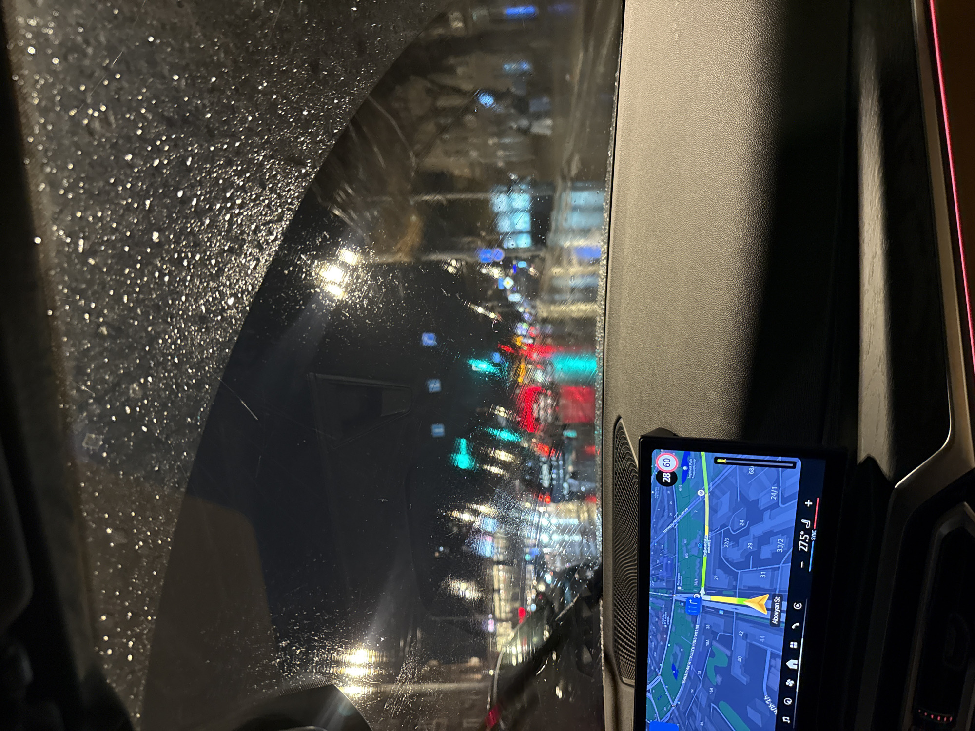The image shows a view from inside a car, looking out through a rain-splattered windshield at a city street at night. The city street is lined with buildings and lit by streetlights and the lights from the buildings. The car's navigation system is visible, displaying a map of the city. The map shows the car's location on Abovyan Street in Yerevan, Armenia. The screen also displays the temperature as 27.5 degrees and a speed limit of 60 km/h.