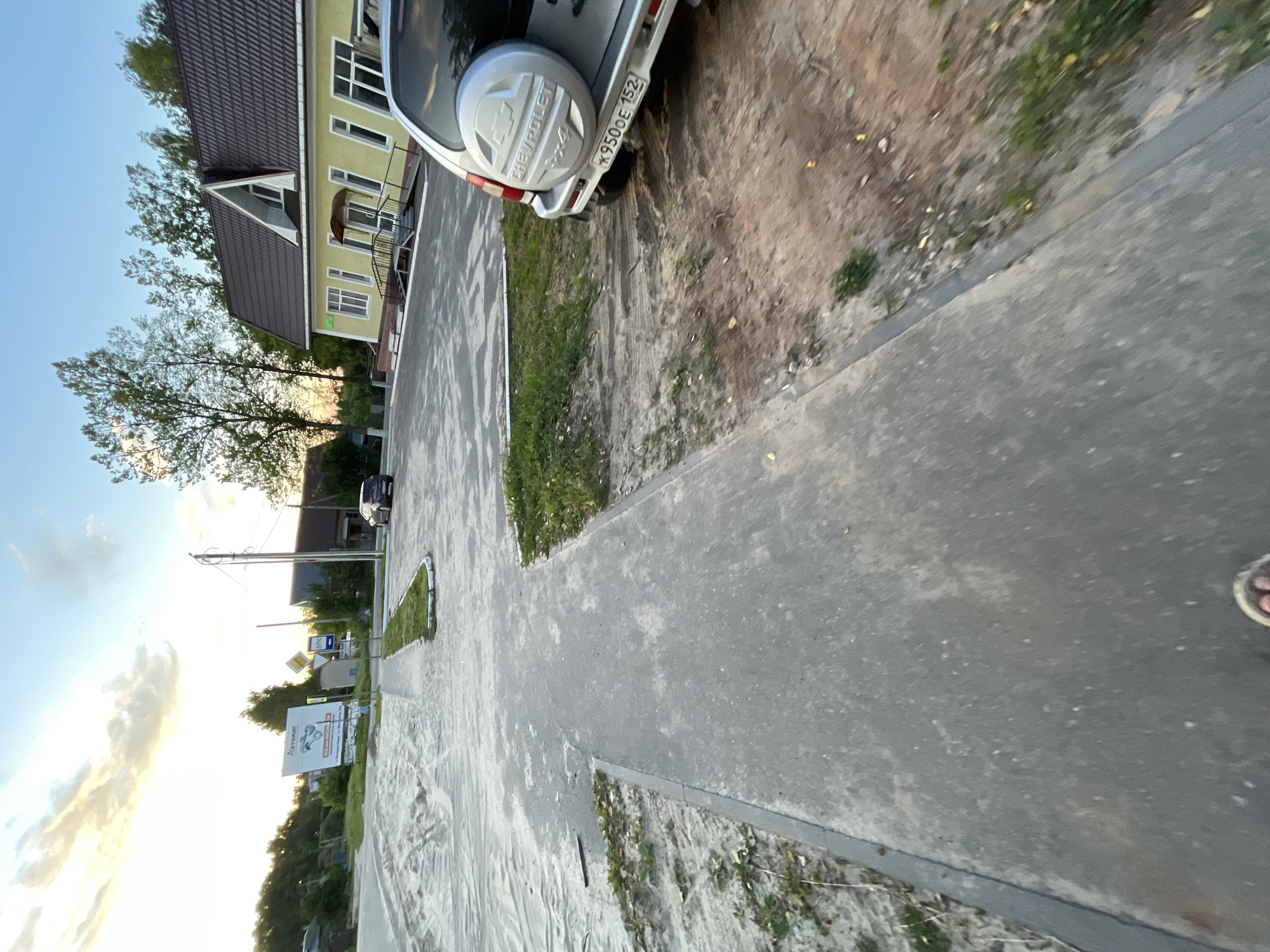 The image captures an outdoor scene in the city of Krasnye Baki, Russia. In the foreground, a paved walkway with a curb is visible, with sandy ground and sparse vegetation to the left. The walkway leads towards a junction. To the right of the walkway, a dirt area with tire tracks is present, bordering a parking lot.
In the mid-ground, a yellow building with a brown roof and white window frames stands prominently. An awning extends over the entrance, which is accessible by a ramp and stairs. Several trees surround the building. A silver Chevrolet Niva SUV is parked in the lot, facing away from the camera, with its spare tire cover visible. The license plate reads "X950OE 152".
Further in the background, a large billboard with Russian text and an image of a car is situated on the left. Several road signs are also visible. A dark-colored car is parked in front of the yellow building. The sky above is a clear blue with a few wispy clouds, and the sun appears to be setting, casting a warm glow. The overall environment suggests a commercial or residential area on the outskirts of the city, possibly near a main road.