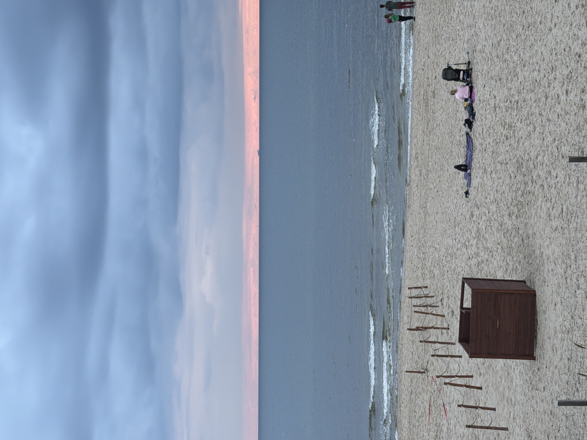 The image depicts a serene beach scene in Międzyzdroje, Poland, under a cloudy, twilight sky. The sea, a calm expanse of blue-grey water, meets the sandy shore where gentle waves lap at the edge. A thin band of pink light is visible on the horizon, separating the sea from the darker sky.
On the beach, a few scattered figures are present. In the foreground, a dark wooden structure, possibly a changing room or information booth, sits on the sand. Further back, a group of people are seen lounging on the beach. One person is seated on a purple mat, facing away from the camera, with a baby stroller beside them. Another person is lying on a dark mat. In the distance, on the right side of the frame, three individuals are standing near the water's edge, their backs to the camera.
A rope and post barrier extends across the sand, creating a pathway. The beach itself is composed of fine, light-colored sand, with subtle undulations and footprints visible. The overall atmosphere is tranquil and peaceful, characteristic of a beach during the transitional hours of dusk.