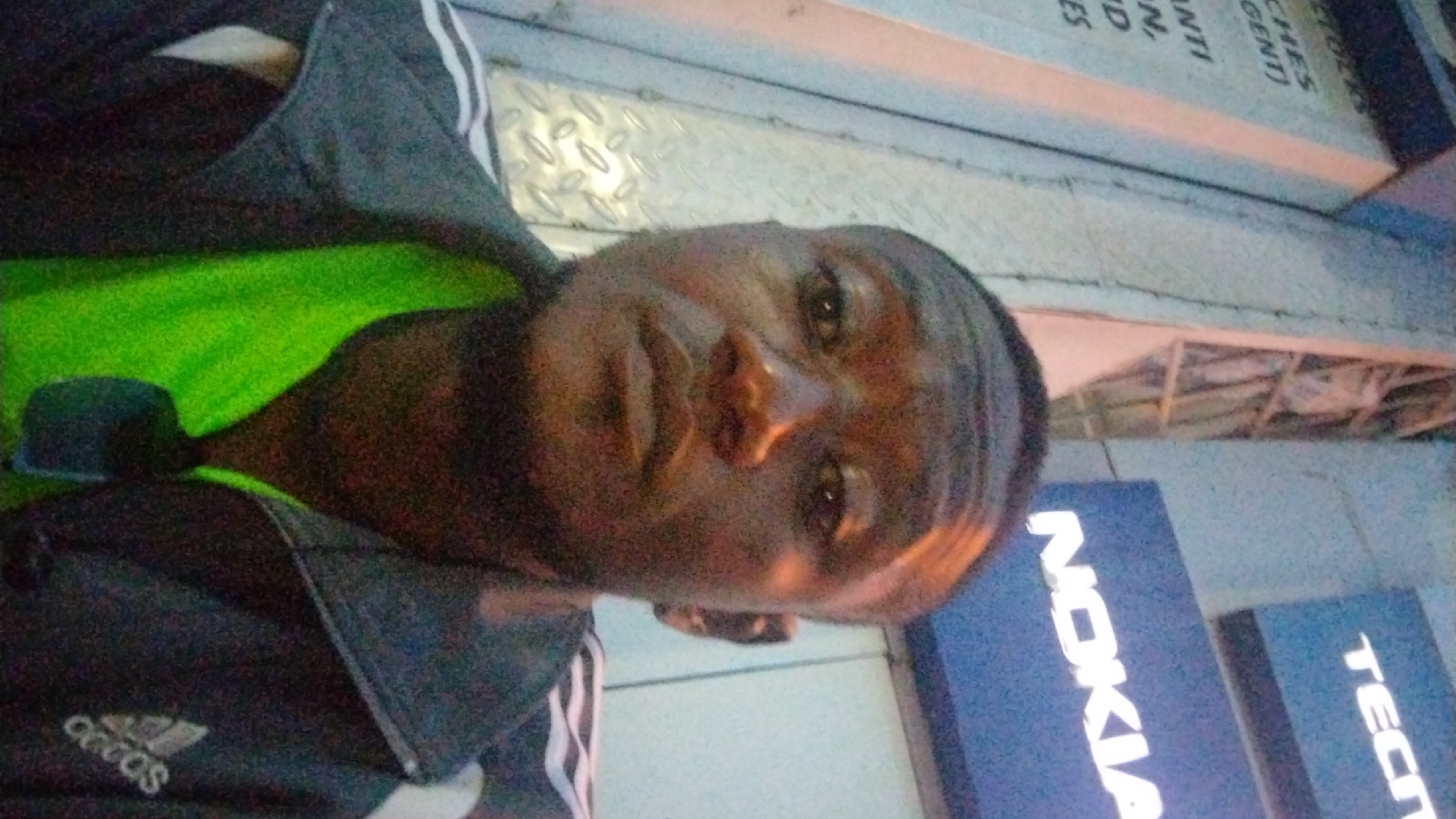 The image, rotated counter-clockwise, captures a dark-skinned individual, appearing to be male, taking a selfie outdoors in what seems to be an urban or commercial setting during the evening or night. The person looks directly at the camera with a neutral to slightly pensive expression.
He is wearing a dark-colored jacket or hoodie that features a white "Adidas" logo on the left chest, layered over a bright lime green shirt, the collar and upper chest of which are visible.
The background suggests a bustling street or shopping area. Immediately behind the person, there's a metallic surface with a diamond plate texture. Further back, several business signs are visible. A large blue sign with the word "NOKIA" in white letters is prominent, alongside another blue sign displaying partially visible white text "TECH". Above these, on a lighter-colored building facade or sign, fragments of text like "...ES", "...NTI", and "...GENT)" can be faintly discerned. The lighting appears artificial, with glows and reflections indicating streetlights or shop illuminations, contributing to the somewhat grainy quality of the photograph, typical of low-light conditions. The scene implies a casual moment captured in a commercial district of Jos, Nigeria.