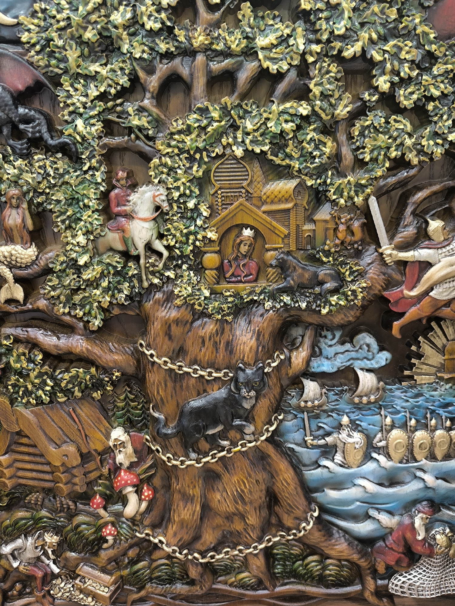 The image shows a highly detailed, carved wooden relief depicting a scene from folklore. A large, gnarled tree dominates the center, with a black cat chained to its trunk. Perched within the branches are various figures and structures. A woman with a crown sits inside a small wooden building. A man on a white horse rides past. Another man with a sword appears to be in combat. Below the tree, a gnome-like figure with a long beard examines mushrooms. To the lower left, a figure in armor kneels beside an open treasure chest. In the background, ships sail on a blue sea, with warriors on board. The overall scene is rich with symbolism and narrative elements, suggesting a story or myth being told through the carving. The image is a close-up, taken from a slight angle, showcasing the texture and depth of the carving. This artwork is not located in Novosibirsk, Russia, but rather appears to be a standalone piece of art.