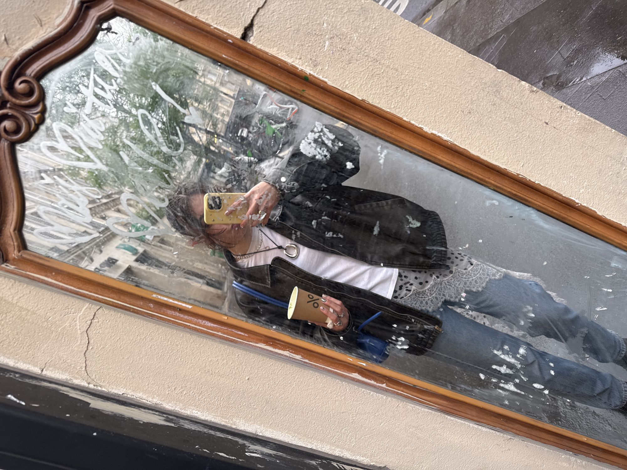 A mirror with an ornate wooden frame is mounted on a beige wall outdoors. The reflection shows a person taking a picture of themselves. The person is wearing a black jacket, a white shirt, and blue jeans with white paint splatter. They are holding a yellow smartphone in their left hand and a brown paper coffee cup in their right. The background visible through the reflection includes a street scene, possibly in Paris, with building facades and trees. There is some graffiti or writing on the mirror itself. The wall on which the mirror is mounted shows signs of wear and cracking.