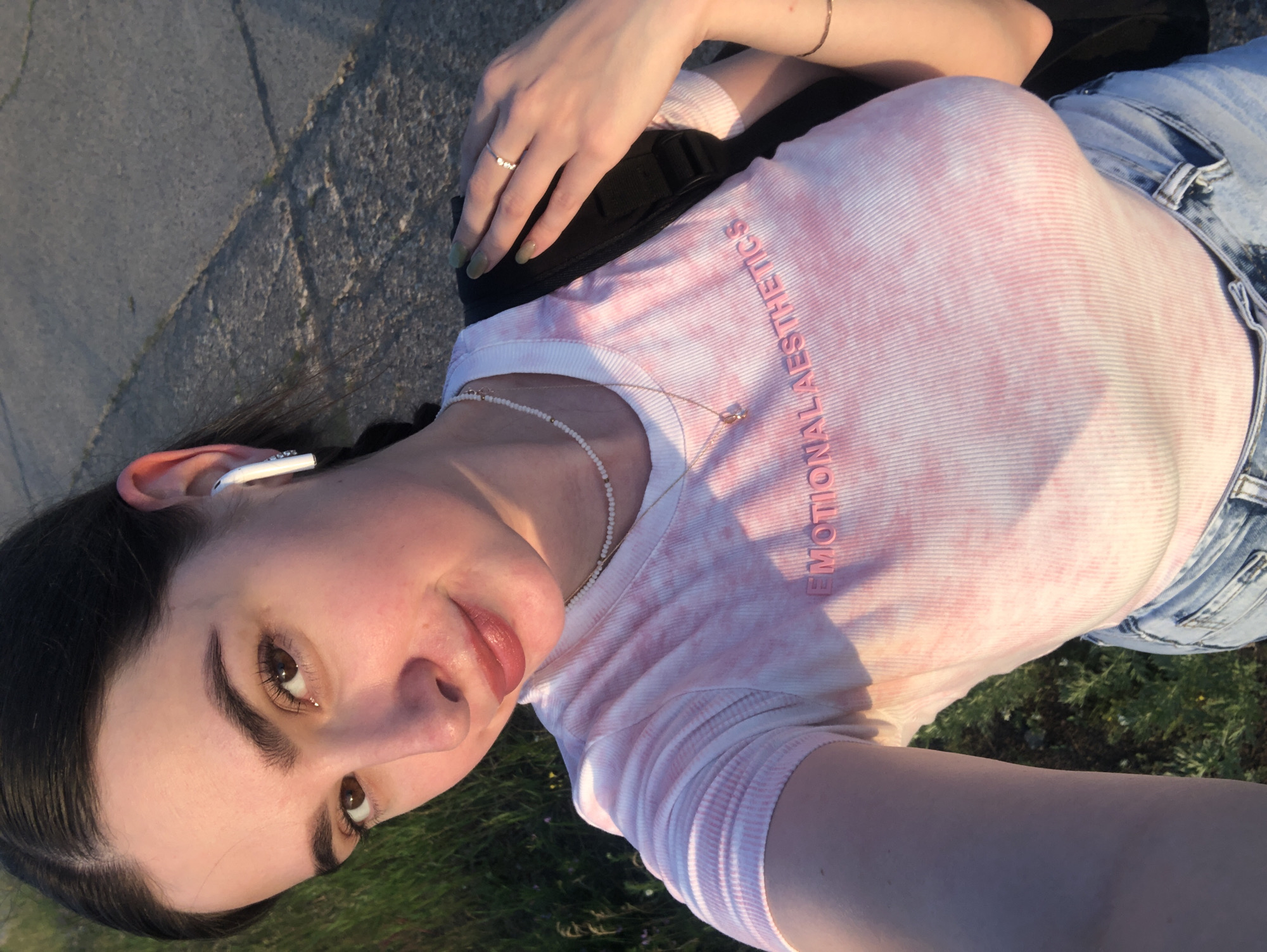 A young woman with dark hair pulled back and a single white earbud in her right ear smiles at the camera. She wears a pink and white tie-dye t-shirt with the words "EMOTIONAL AESTHETICS" printed in pink on the chest. She also wears a delicate pearl necklace and a gold chain with a pendant. A thin gold bracelet is visible on her right wrist. Her left hand, adorned with a simple ring, rests on a black backpack strap. She is wearing light blue denim jeans. The background shows a paved surface and green grass with some small yellow flowers, suggesting an outdoor setting possibly in the city of Chaikovsky, Russia, perhaps along a sidewalk or park path during sunset, as indicated by the warm lighting.