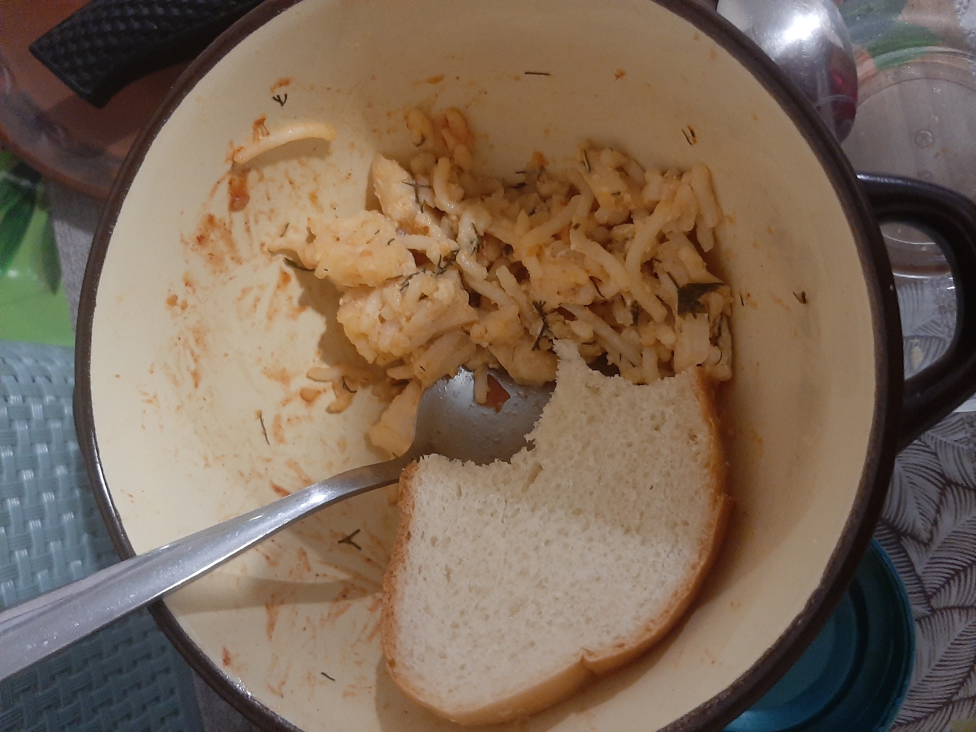 An overhead, close-up shot shows a light yellow, well-worn bowl containing a small portion of food and a slice of white bread. The food appears to be a mixture of rice or pasta with small pieces of what might be tomato and herbs. A silver spoon rests on the food, with its handle extending out of the frame. The slice of bread, which has a soft, white interior and a slightly darker crust, is positioned to the left of the food, partially covering it. The bowl itself has a dark brown rim and is placed on a surface with a patterned cloth underneath. In the background, a green object and a dark-colored object are visible. The scene is lit from above, casting shadows within the bowl.